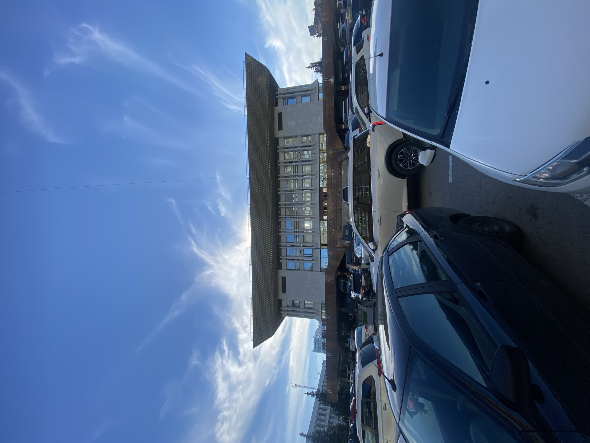 The image depicts the Ufa Congress Hall Toratau, a modern building with a distinctive cantilevered roof, in Ufa, Russia. The building is situated in a large outdoor parking lot filled with numerous cars. The sky above is bright blue with wispy white clouds. The perspective is from ground level, looking up at the building and the sky, with several cars in the foreground obscuring the view of the lower part of the building. The lighting suggests it is daytime, possibly late afternoon due to the angle of the sun, which is partially obscured by the building's roof, casting a bright glow. No people are visible in the frame. The environment appears to be an urban setting.