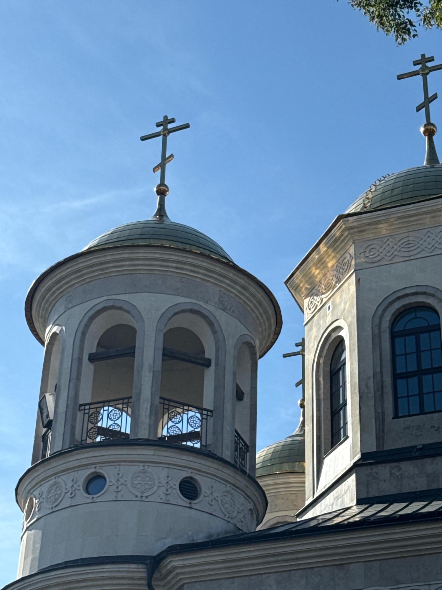 This image captures a close-up, low-angle view of two Eastern Orthodox church domes against a clear blue sky. The left dome is part of a cylindrical bell tower, which features arched openings with decorative metal railings visible behind them. The tower has a rounded base adorned with circular windows and intricate carvings. To the right, a second, larger dome rises from a square structure. This structure has a tall, arched window with dark panes and decorative stonework along its upper edge. Both domes are capped with multi-tiered Orthodox crosses, with the one on the left appearing to be a more traditional eight-pointed cross. The building materials are primarily light-colored stone or stucco, with golden accents on the domes and crosses. The overall scene is bathed in sunlight, casting subtle shadows and highlighting the architectural details. The image is likely of a church in Zvenigorod, Russia, given the architectural style commonly found in Russian Orthodox churches.