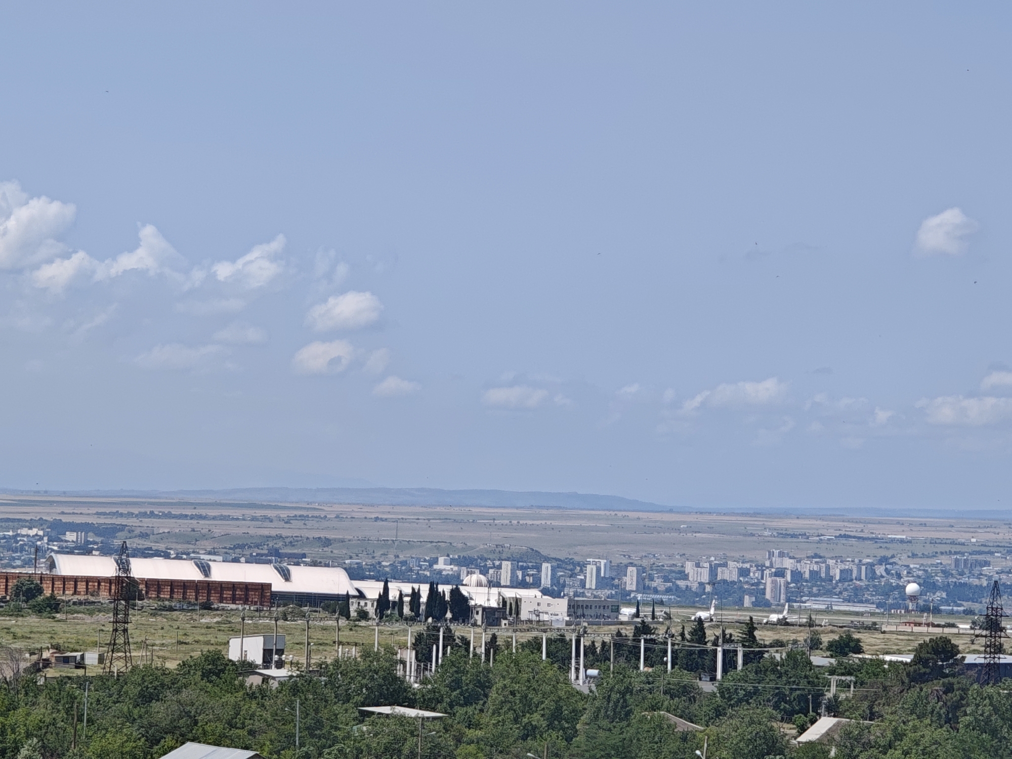 This is a wide-angle, eye-level shot of the Tbilisi, Georgia skyline on a clear day. The sky is a bright, pale blue with scattered white clouds. In the foreground, lush green trees fill the lower portion of the frame. Behind the trees, a large, elongated building with a white, arched roof is visible, flanked by rows of rust-colored structures. Several electrical towers and smaller buildings are interspersed within the landscape. Further back, a sprawling city unfolds with numerous residential and commercial buildings, including some taller structures. An airport runway is visible in the middle ground, with several airplanes on the tarmac. A large white dome-shaped structure, possibly a weather radar or communication antenna, stands near the airport. The distant horizon is characterized by a hazy, flat landscape with rolling hills. The overall impression is one of urban development within a vast, open geographical area.