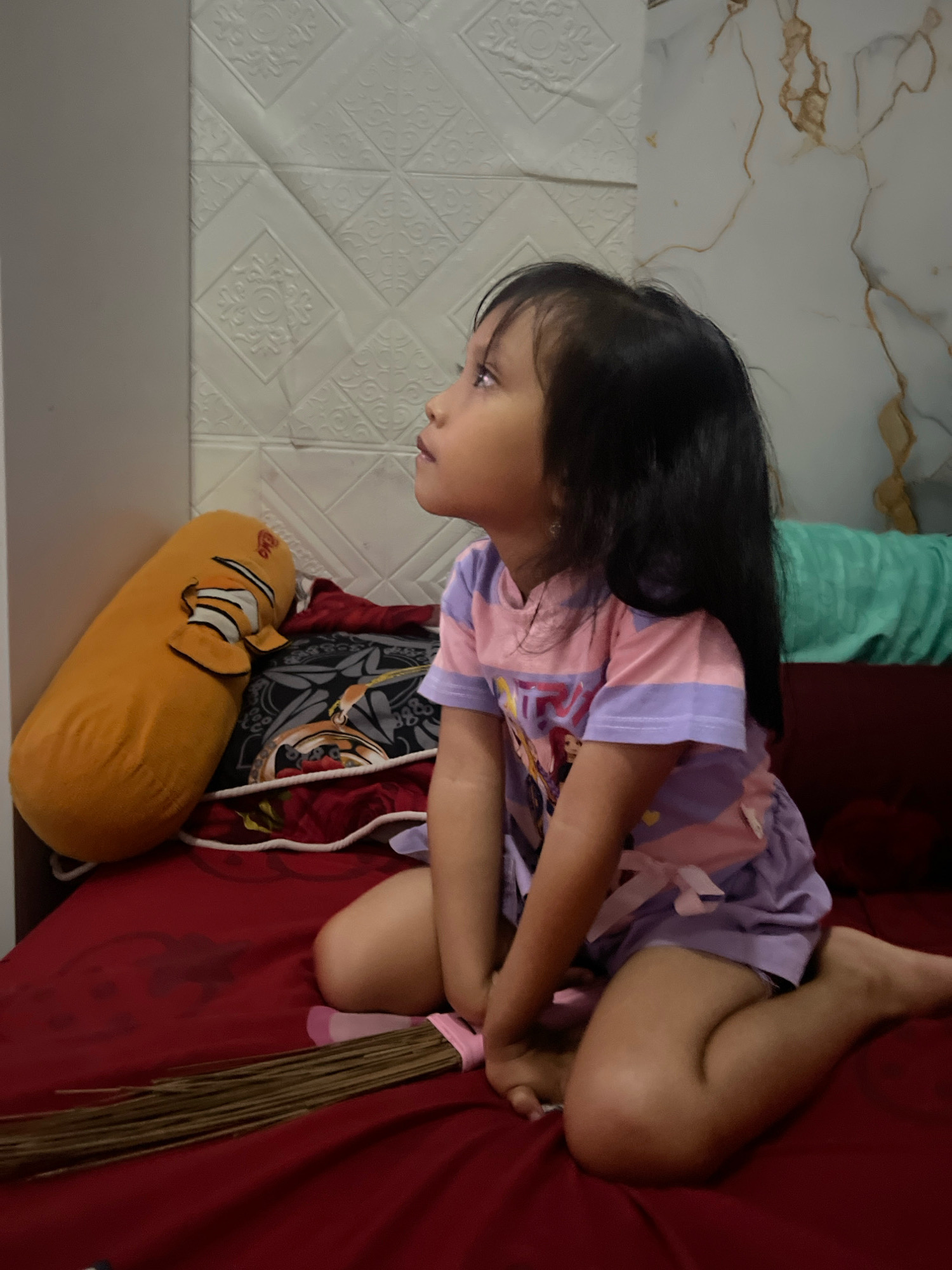 A young girl with long, dark hair sits on a bed with a deep red comforter. She is wearing a short-sleeved shirt with horizontal stripes of pink and lavender, and coordinating lavender shorts. Her arms are resting on her knees, and she is looking upwards and to the left. Her skin is light brown.
To her left, a large, bright orange bolster pillow with a Nemo character printed on it is leaning against a textured white wall panel. Behind the girl, a dark patterned pillow and a green blanket are visible. In front of the girl, resting on the red comforter, is a broom made of dried brown bristles, tied with a pink ribbon. The room appears to be indoors, likely a bedroom, with a patterned wallpaper or wall covering behind the bed. The lighting suggests it is daytime.