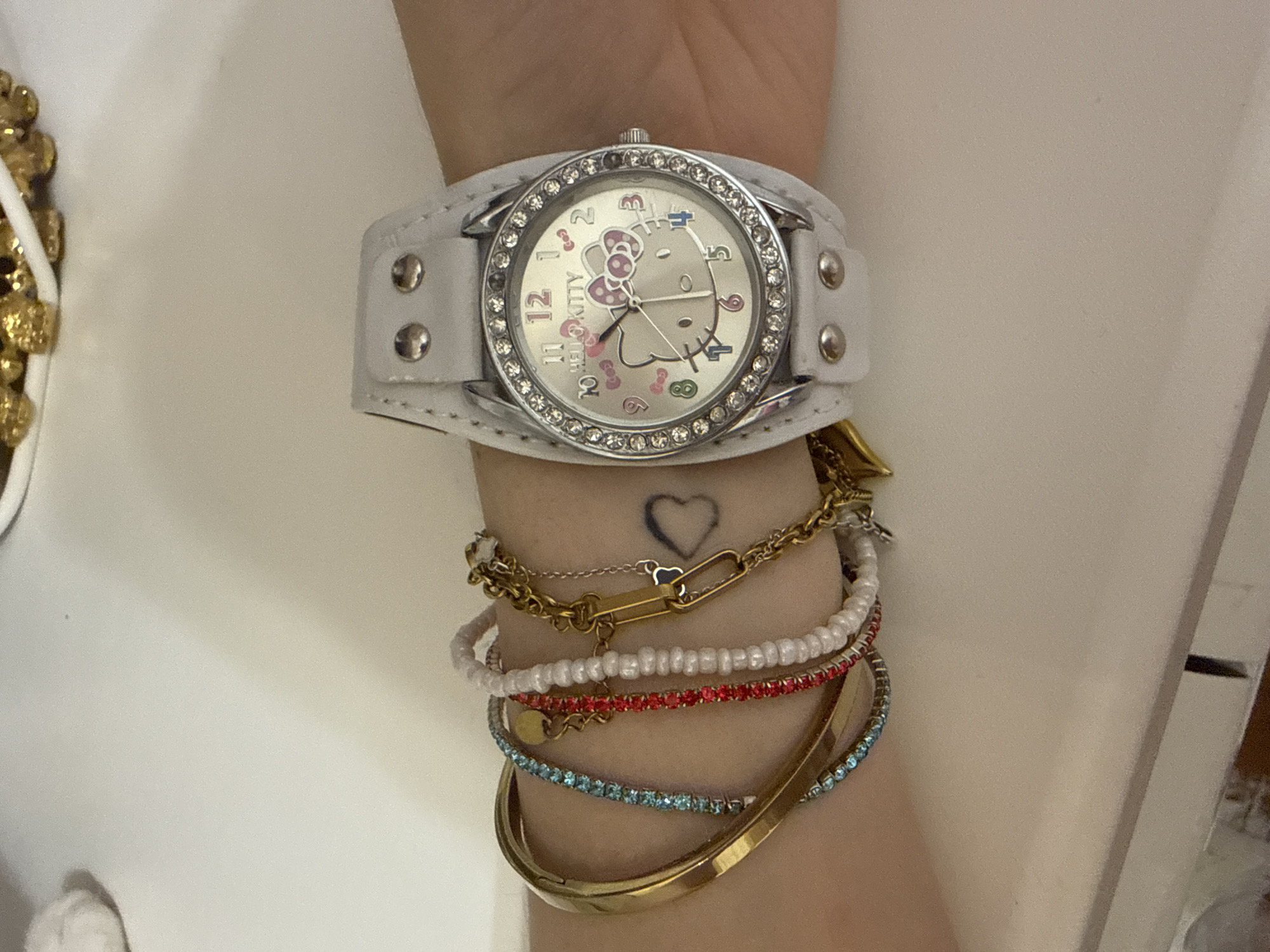 A close-up shot shows a person's left wrist adorned with multiple bracelets and a white Hello Kitty watch. The bracelets include a thick gold bangle, a thin bangle with blue rhinestones, a bangle with red rhinestones, a strand of small white beads, and a gold chain bracelet with an oblong link and a small gold charm. The white watch has a silver-toned bezel encrusted with rhinestones, and the watch face features a silver background with the Hello Kitty logo and an illustration of Hello Kitty. The watch band is white leather with silver studs. A small heart tattoo is visible on the person's skin beneath the bracelets. The background is a plain, light-colored surface, likely a table.