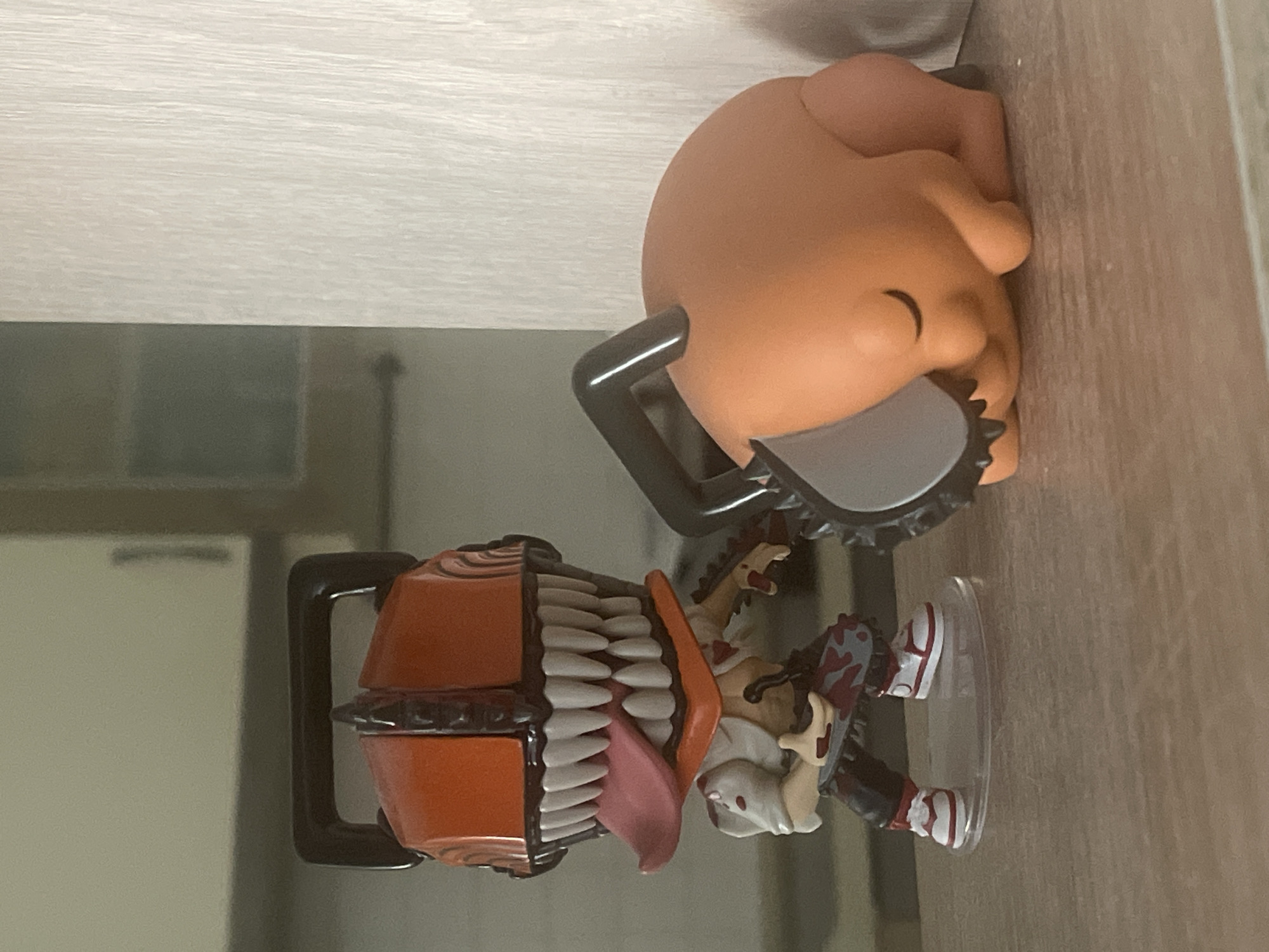 In a dimly lit indoor setting, likely a shelf or countertop, two figurines are positioned. The figurine on the left is a stylized character with an orange, chainsaw-like head featuring sharp white teeth and a pink tongue protruding. It wears a white shirt with blood splatters, dark pants with a chain saw design, and white sneakers with red accents. The figurine stands on a clear plastic base. To the right, a smaller, orange, round figurine is curled up and appears to be sleeping. It has closed eyes and its body resembles a smooth, rounded form. A dark handle and part of a chainsaw blade are positioned on top of this figurine. The background is blurred, showing hints of cabinetry and a textured, light-colored vertical surface. The location is not specified in the image.