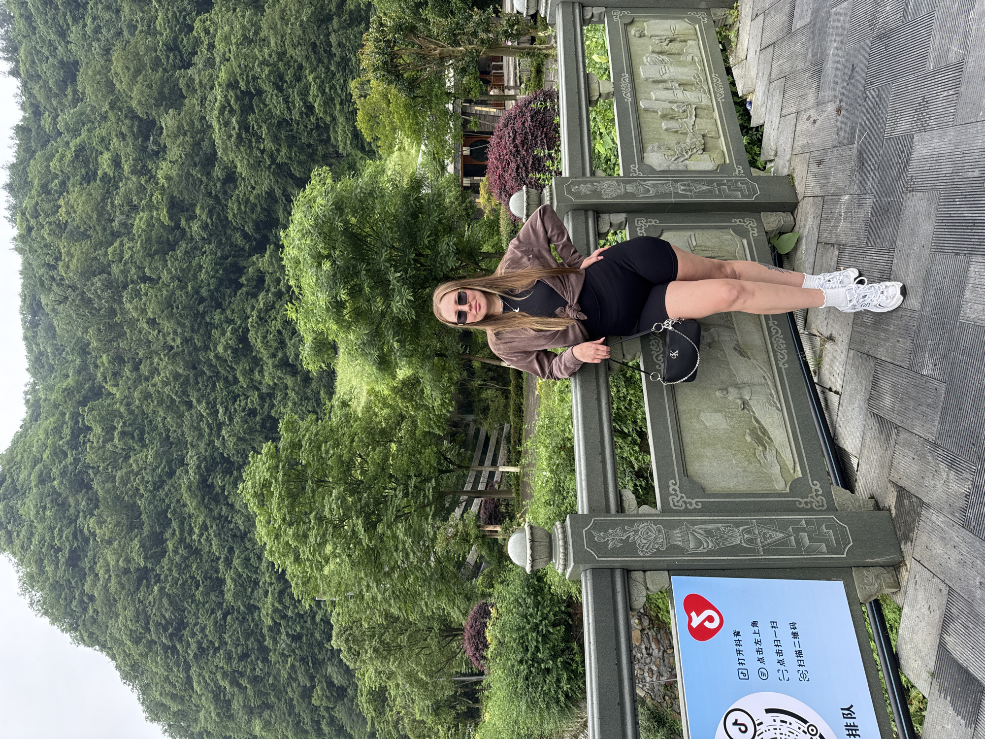 A woman with long blonde hair, wearing a black top, black shorts, a brown jacket, white socks, and white sneakers, is lying on a stone bridge in Jundiping, China. She is wearing sunglasses and has a black purse with a silver chain next to her. The bridge has ornate carvings and a sign with Chinese characters and a QR code. Behind the bridge are trees, and a steep, green, heavily wooded hillside.