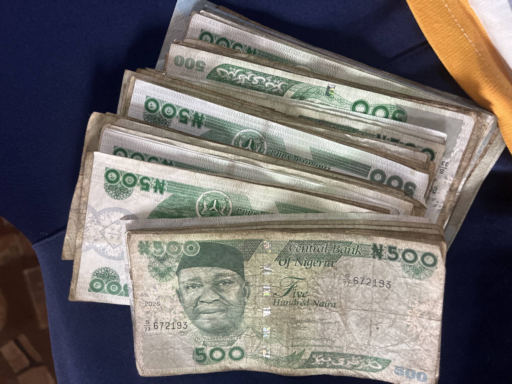 A close-up, overhead shot shows a stack of Nigerian 500 Naira banknotes fanned out on a dark blue fabric, possibly clothing or upholstery. The banknotes are predominantly green and white, featuring the portrait of a man on the left and numerical denominations. Some of the notes are worn and creased, indicating use. The lighting suggests an indoor setting. The exact location of Nkpor, Nigeria cannot be determined from the image alone.