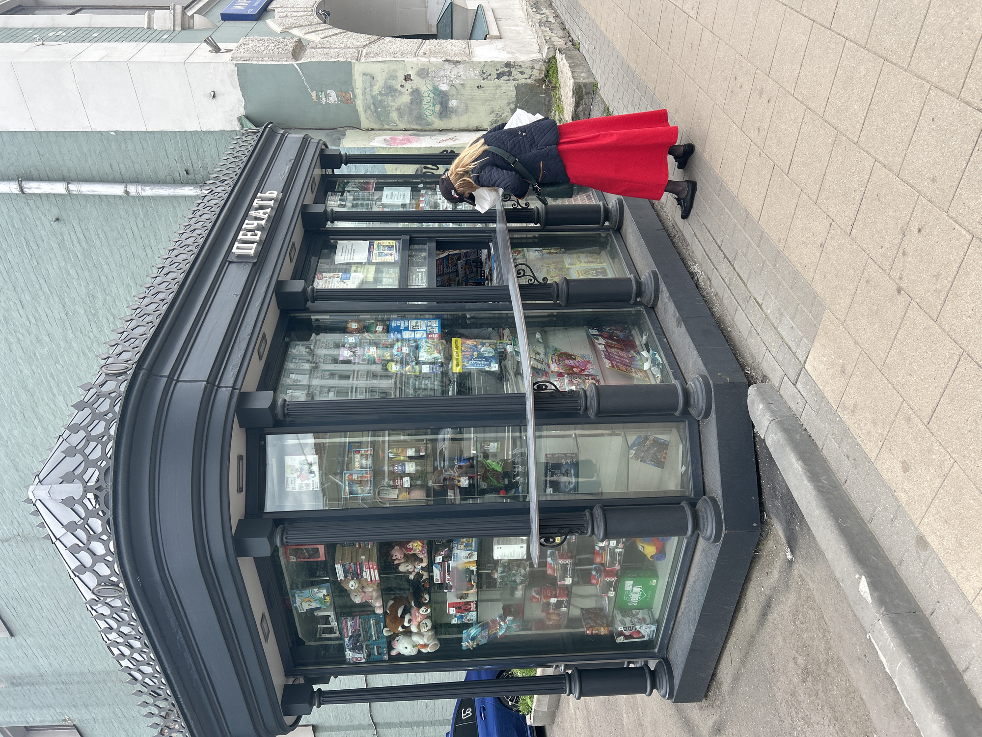 The image displays an urban street scene featuring a dark-colored newsstand or kiosk with a person interacting with it. The kiosk is rectangular with a rounded corner, constructed from dark metal framing extensive glass display panels. These panels reveal various items for sale, including magazines, newspapers, small toys, and other merchandise. An ornate, possibly metallic, decorative roof structure with a repeating scalloped pattern sits atop the kiosk. The Cyrillic word "ПЕЧАТЬ" (PECHAT', meaning "Press" or "Print") is prominently displayed in raised, metallic letters on the kiosk's upper frame.
A person with light-colored hair, wearing a long red skirt, a dark quilted jacket, and dark shoes, is leaning into the kiosk, appearing to browse or make a purchase. A white bag or item is held near their face, and a dark shoulder bag hangs from their shoulder.
The kiosk is situated on a light-colored paved sidewalk made of square tiles. A gray concrete curb runs alongside, separating the sidewalk from a darker, possibly asphalted, street surface. In the background, a building with a light blue-green painted lower facade and a lighter, weathered upper section is visible, showing signs of wear and some faint markings. A small portion of a blue car is visible at the base of the kiosk. The lighting suggests daylight, possibly under an overcast sky. The scene depicts a typical street vendor interaction in an urban environment, consistent with a location like Moscow, Russia.