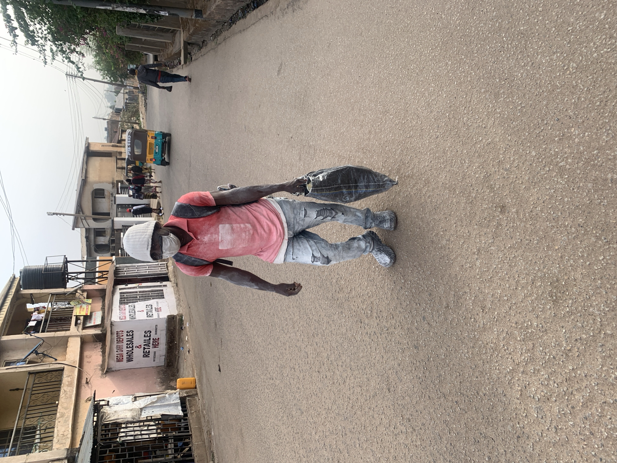 A man in a red shirt and light-colored pants and shoes walks on a paved street carrying a dark, heavy bag. His skin has light splotches. A white cap and face mask obscure his head and face. In the background, a three-wheeled vehicle travels down the street. A sign for "MEGA GARI DEPOTS WHOLESALES & RETAILERS HERE" is visible on a nearby building. The setting appears to be a street in Jos, Nigeria.