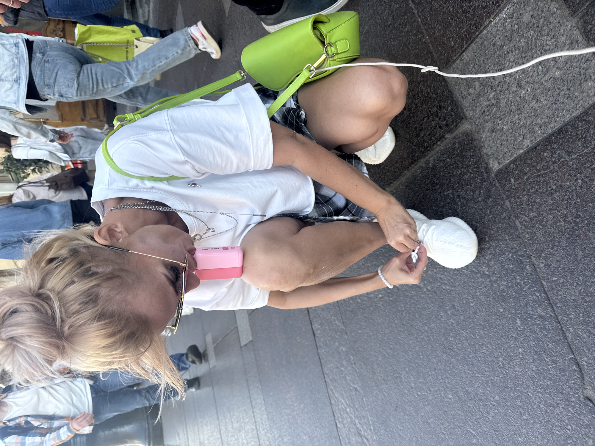 The image shows a woman crouching down to tie her white sneaker. She has blonde hair pulled up in a messy bun and is wearing glasses, a white t-shirt, and a plaid skirt. A bright green purse is slung over her shoulder. She is holding a pink object in her left hand near her face. She is on a stone-paved sidewalk, likely in a public area, with other people in the background. The setting appears to be a sunny day. The architectural details of the buildings in the background suggest a European city, possibly Saint Petersburg, Russia, given the presence of a style of paving stones and architectural elements commonly seen in that city.