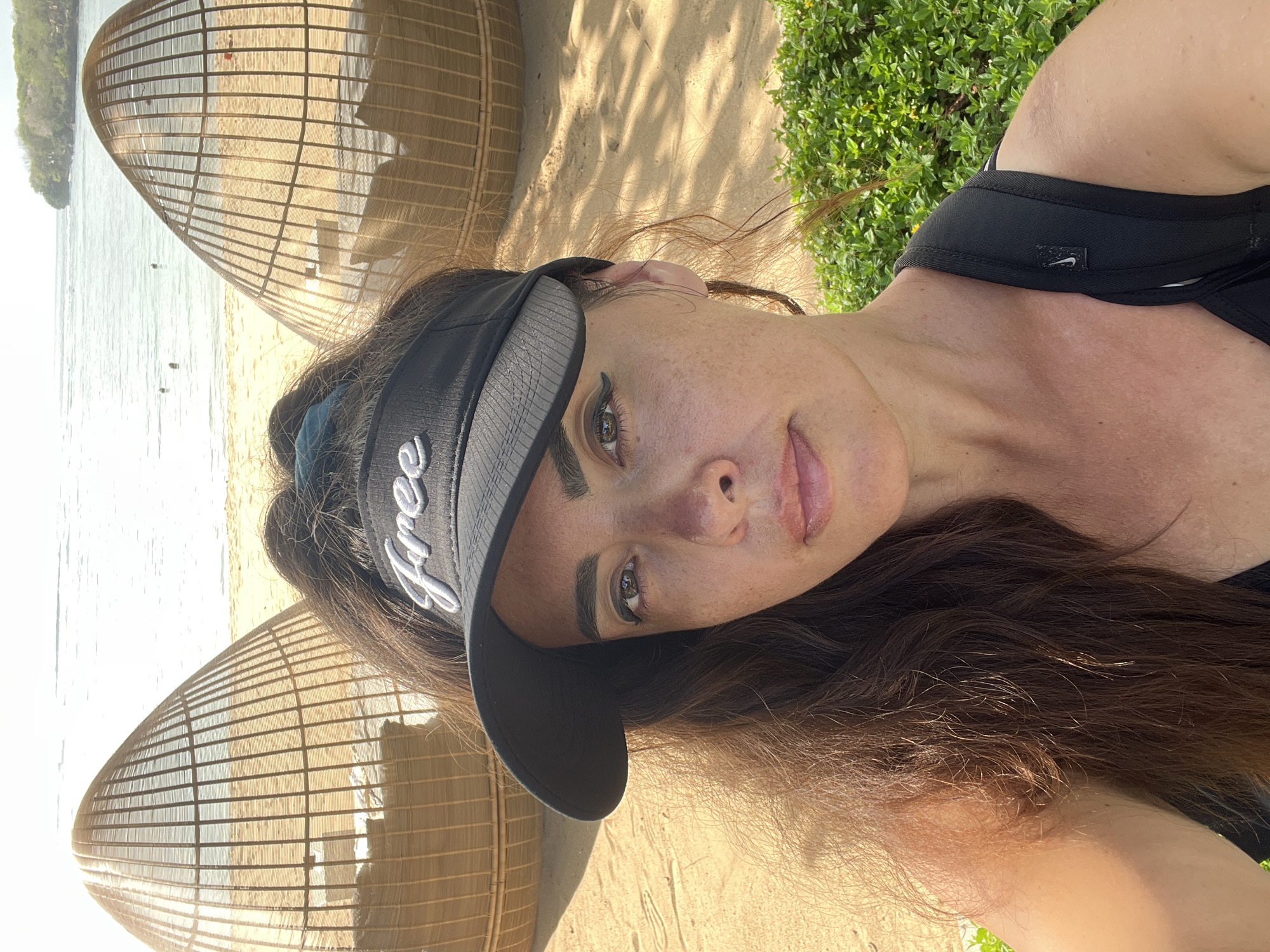 An outdoor, eye-level, medium shot captures a woman with long, dark brown hair and a black visor with the word "Free" embroidered in white, posing for a selfie on a sandy beach. She is wearing a black top. Her skin is lightly tanned and speckled with freckles. Her hair is pulled back and cascades over her left shoulder. In the background, two large, woven, egg-shaped lounge chairs are visible on the sand. Beyond the chairs, the ocean stretches out under a bright, sunny sky, with a distant green-covered cliff on the right horizon. A line of green bushes borders the lower right of the frame. The scene suggests a tropical beach resort setting in Kuta Selatan, Indonesia.