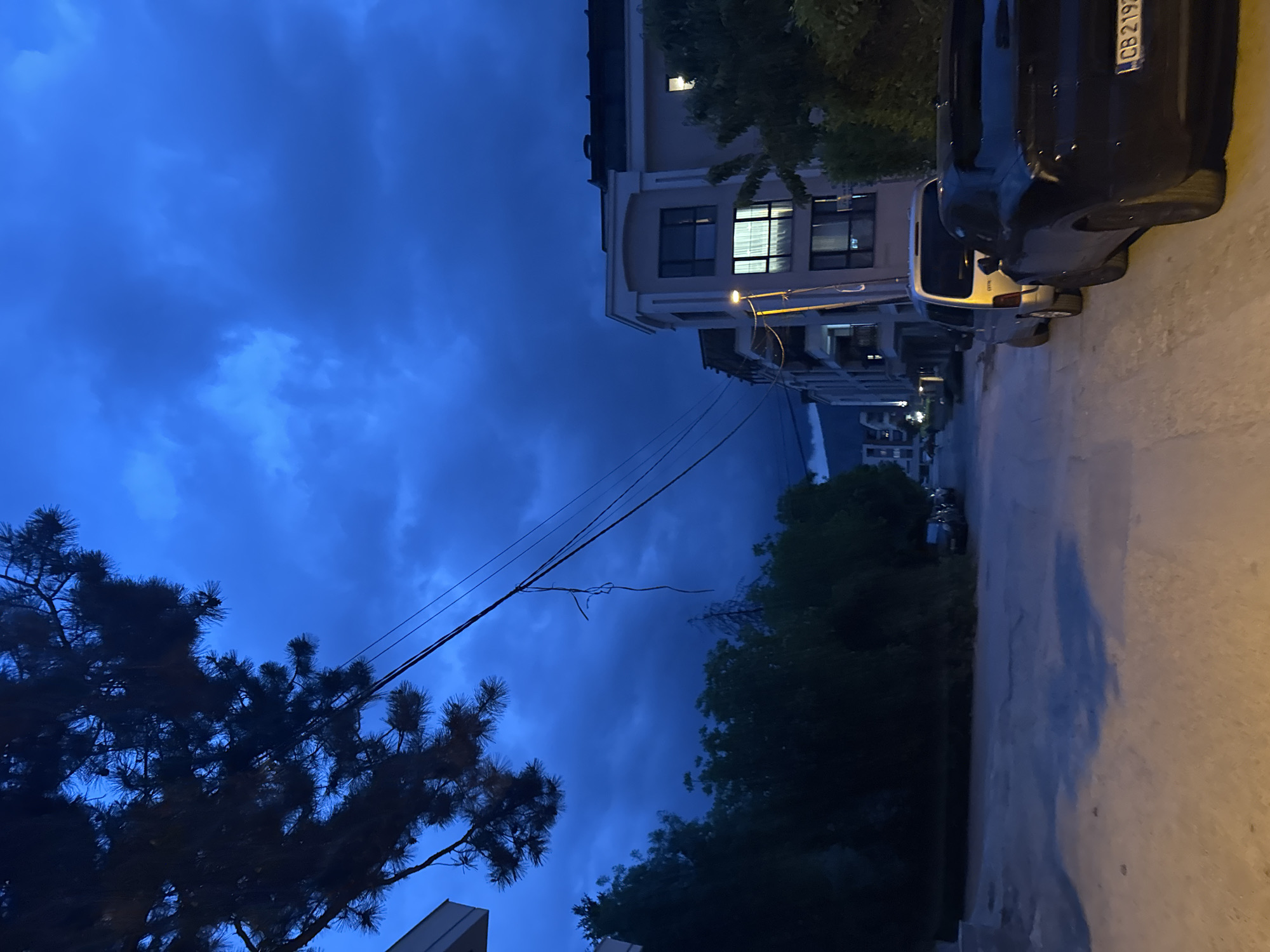 The image depicts a nighttime scene in Sofia, Bulgaria. The sky is a deep blue, filled with heavy clouds. Power lines stretch across the sky. Below, there is a row of buildings, with some windows lit. A street lamp illuminates the area. Cars are parked along the side of the road. Trees are visible, some in silhouette against the dark sky. A building with "CB 219" on a license plate can be seen. The overall mood is somber due to the dim light and overcast sky.