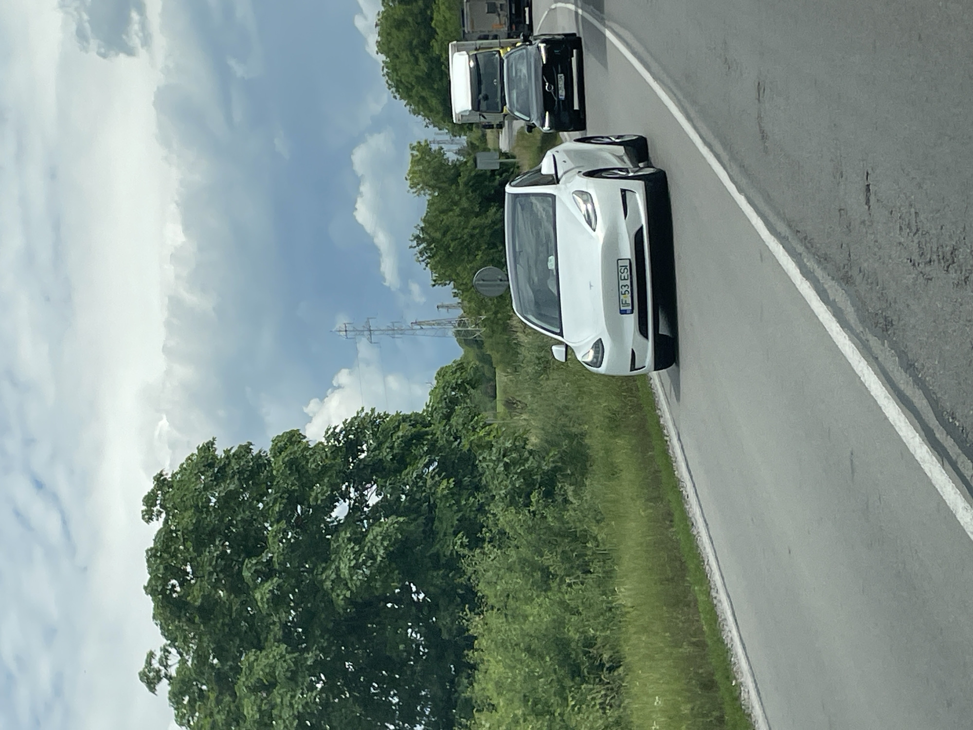 The image depicts a white Tesla Model Y with license plate "IF 53 ESI" driving on a two-lane asphalt road. Adjacent to the car is a grassy shoulder and lush green trees. Further ahead, a black Volvo and a white truck are also driving on the road. Power lines are visible in the background, and the sky is partly cloudy. There is no verifiable evidence to support a location in Getsovo, Bulgaria.