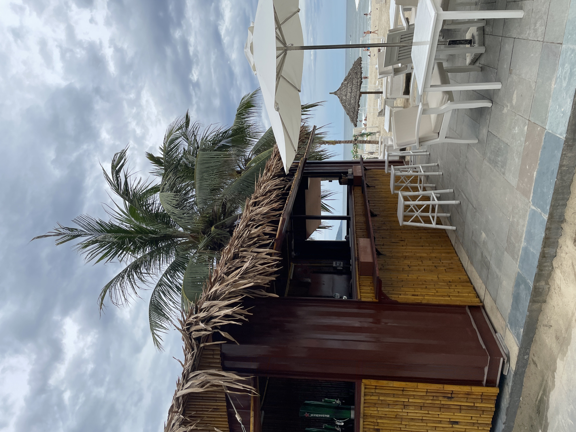 The image depicts an outdoor bar and seating area on a beach, likely in the city of Đà Nẵng, Vietnam, given the style of the beach bar and common tourist destinations in the region. The scene is set under a cloudy sky, with palm trees swaying in the background.
In the foreground, a rustic beach bar with a thatched roof is visible. The bar itself is constructed from dark brown corrugated metal and bamboo. On the left side of the bar, two green beer taps with the Heineken logo are prominent. A wooden counter runs along the front of the bar, with several white bar stools placed in front of it.
Adjacent to the bar, a seating area is set up with white tables and chairs. A large white umbrella provides shade over one of the tables. Further back, on the sandy beach, several other thatched umbrellas and lounge chairs are visible.
The beach is populated by a few individuals, some walking or standing near the water, and others relaxing on the beach. The ocean stretches to the horizon, with a faint outline of land visible in the distance. The overall atmosphere suggests a relaxed, tropical beach setting, despite the overcast weather.