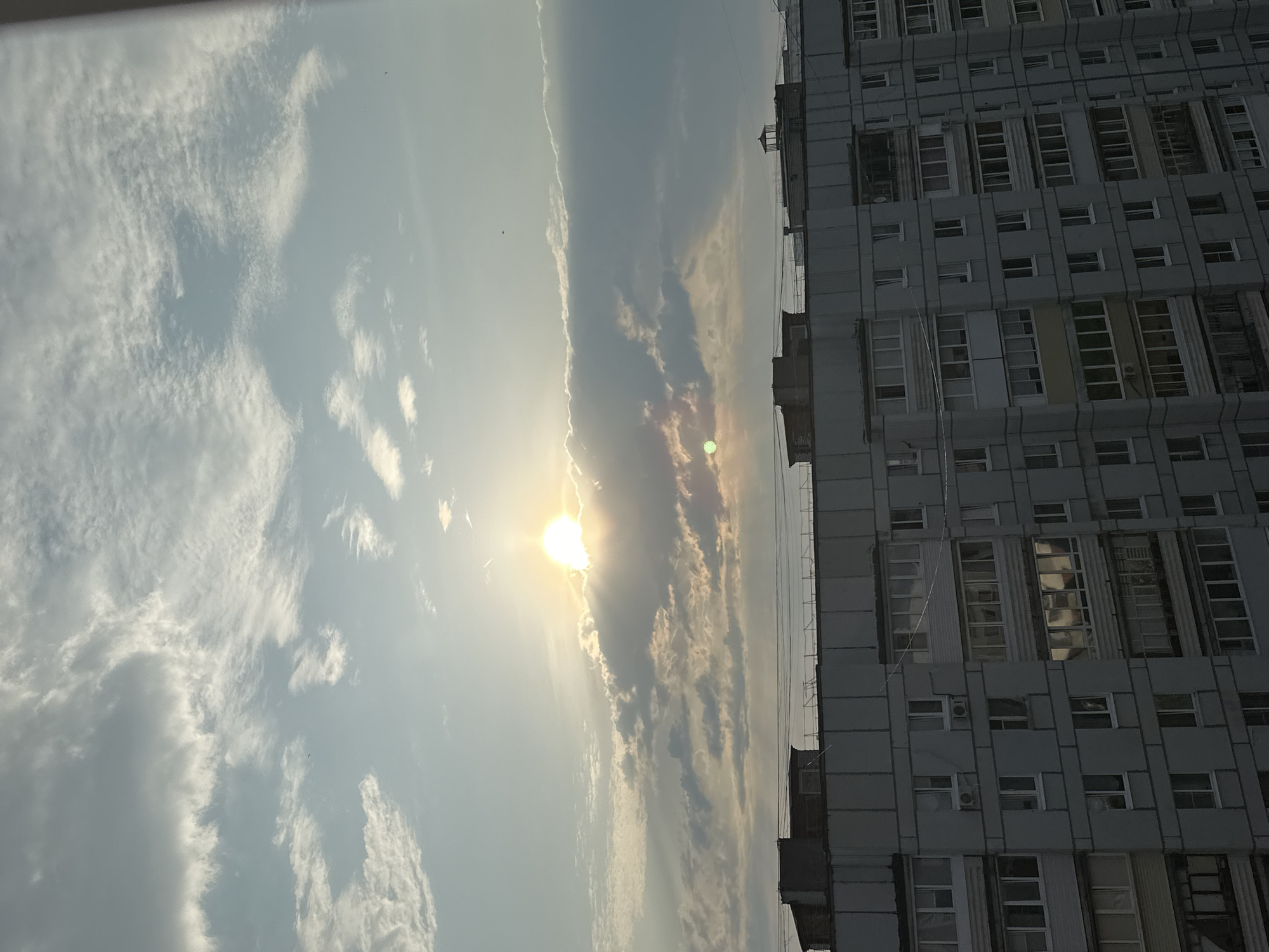 The image captures a view of a residential building against a sky filled with clouds and the sun. The building is a multi-story apartment complex, likely constructed from concrete panels, with numerous windows and balconies. The sunlight is bright and diffused by the clouds, creating a soft illumination across the scene. The setting appears to be urban, with the building dominating the lower portion of the frame and the sky taking up the upper half. The context provided indicates this scene is located in Chaikovsky, Russia.
