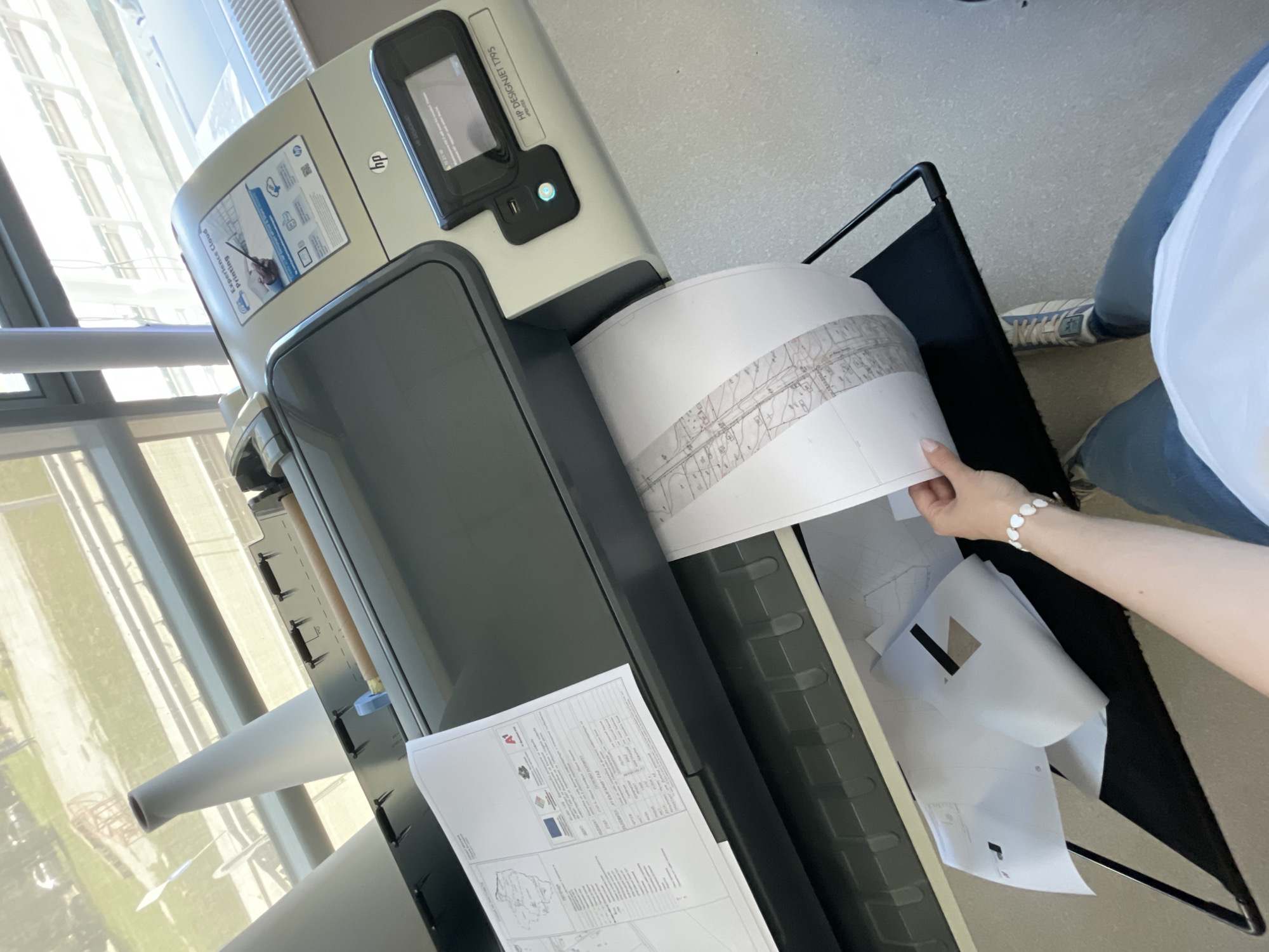 The image depicts an HP DesignJet T795 printer in an indoor setting, possibly an office, located in Sofia, Bulgaria. The printer is shown dispensing printed maps or technical drawings into a black fabric catch basket. A person's arm is visible, with a wrist adorned with a white bracelet, reaching for a rolled-up map emerging from the printer. The background includes a large window and an outdoor view of what appears to be the city of Sofia. Additional printed papers are visible within the catch basket. The setting appears to be well-lit and clean.