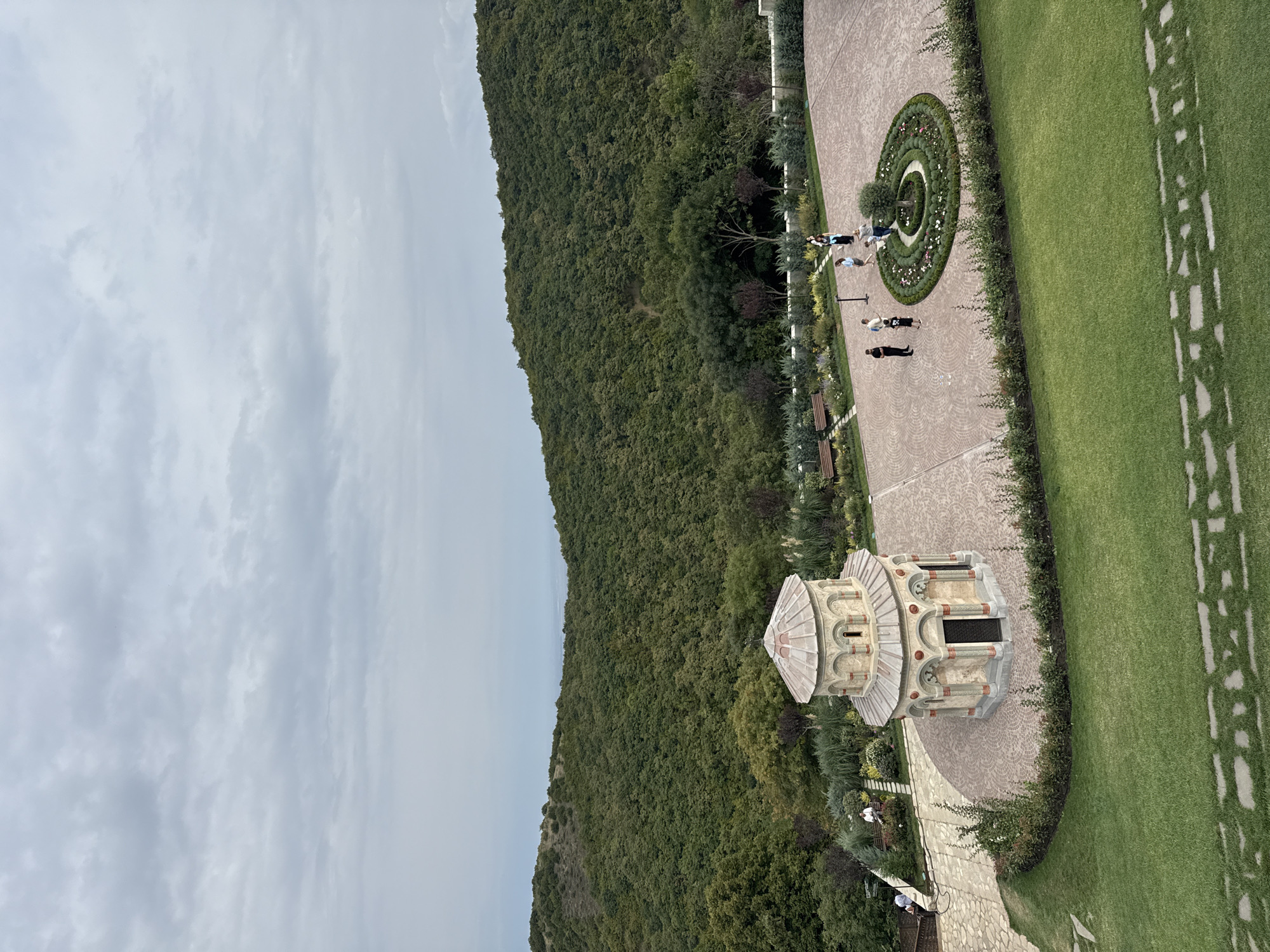 The image depicts a view of the Bodbe Monastery complex in Sighnaghi, Georgia, on an overcast day. In the foreground, a well-maintained lawn with decorative stone pathways leads to a paved courtyard. The courtyard features a small, ornate chapel with a domed roof, surrounded by a circular flower bed with a small tree at its center. Several individuals are visible in the courtyard, some walking and others standing, appearing to be visitors or monks. Beyond the courtyard, the landscape transitions into a lush, green hillside densely covered with trees, characteristic of the Kakheti region. The sky is filled with gray clouds, suggesting a cool or possibly rainy day. The overall scene conveys a sense of tranquility and spiritual significance, integrated with the natural beauty of the Georgian countryside.