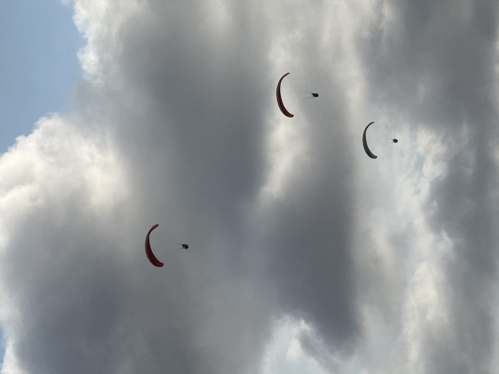 Three paragliders are captured mid-flight against a dramatic sky filled with dark, voluminous clouds and patches of blue sky. The paragliders are descending at varying altitudes. The topmost paraglider has a predominantly red canopy, while the middle one has a red and black canopy. The lowest paraglider's canopy is a dark gray. All paragliders appear to be alone and are not interacting with each other. The scene suggests an outdoor recreational activity, possibly in the vicinity of a popular tourist destination given the context of Kuta Selatan, Indonesia, which is known for its beaches and adventure sports. The lighting is diffused due to the cloud cover, creating a somewhat moody atmosphere.
