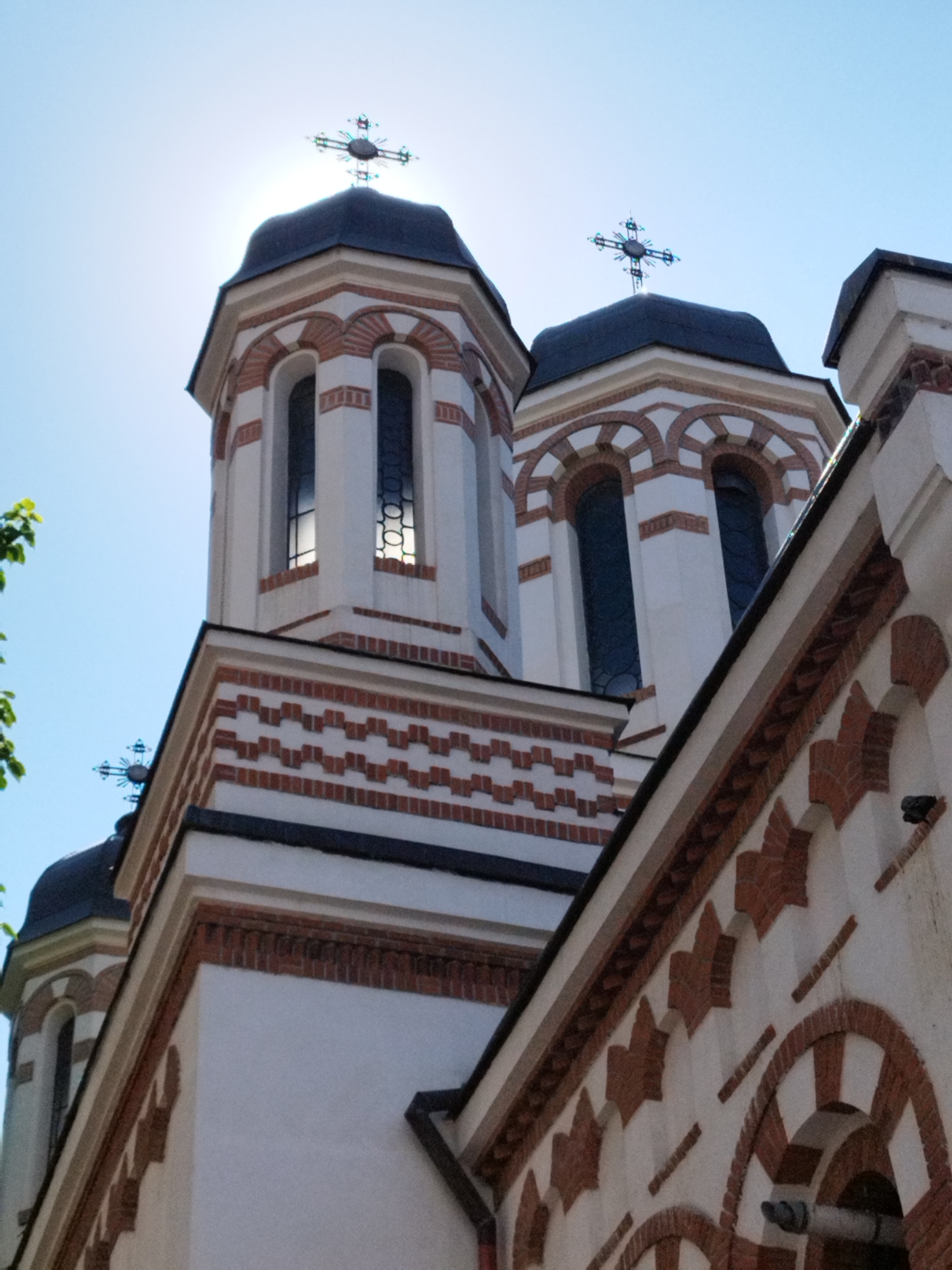 This is a low-angle, medium shot of the exterior of a church in Bucharest, Romania, under a clear blue sky. The sunlight is strong, creating a lens flare behind the cross on the highest tower. The church features white walls with decorative red brickwork in geometric patterns and arched window surrounds. Two prominent towers with dark roofs are visible, topped with ornate crosses. In the foreground and to the left, green leaves from a tree are partially in frame. The architecture suggests a Byzantine or Orthodox style. The image is focused on the upper portions of the church structure.