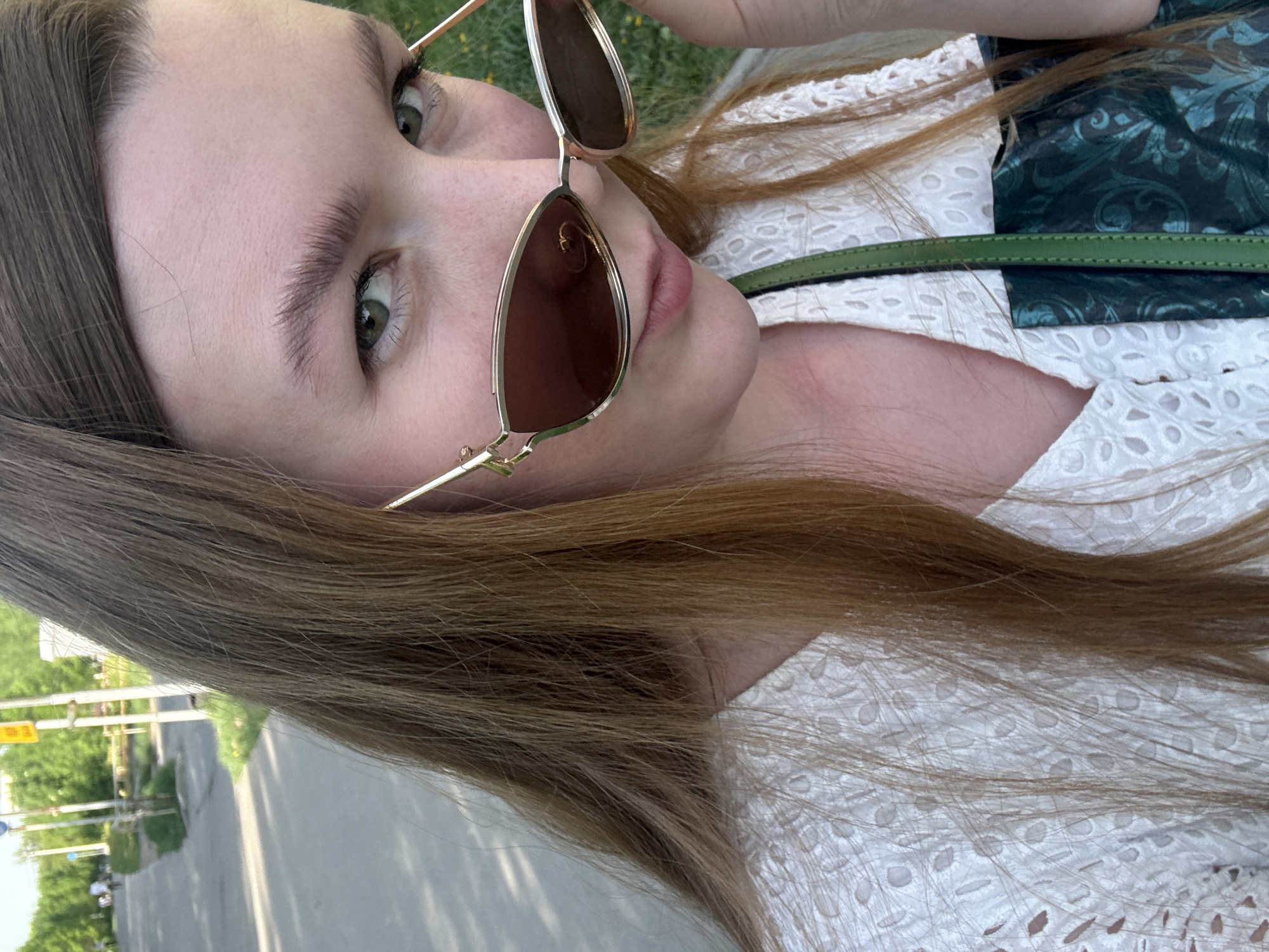 A young woman is depicted at an angle, wearing sunglasses and a white eyelet top with a dark green garment. The background appears to be an urban street scene, possibly near Izhevsk, Russia, showing a pedestrian path and street furniture.