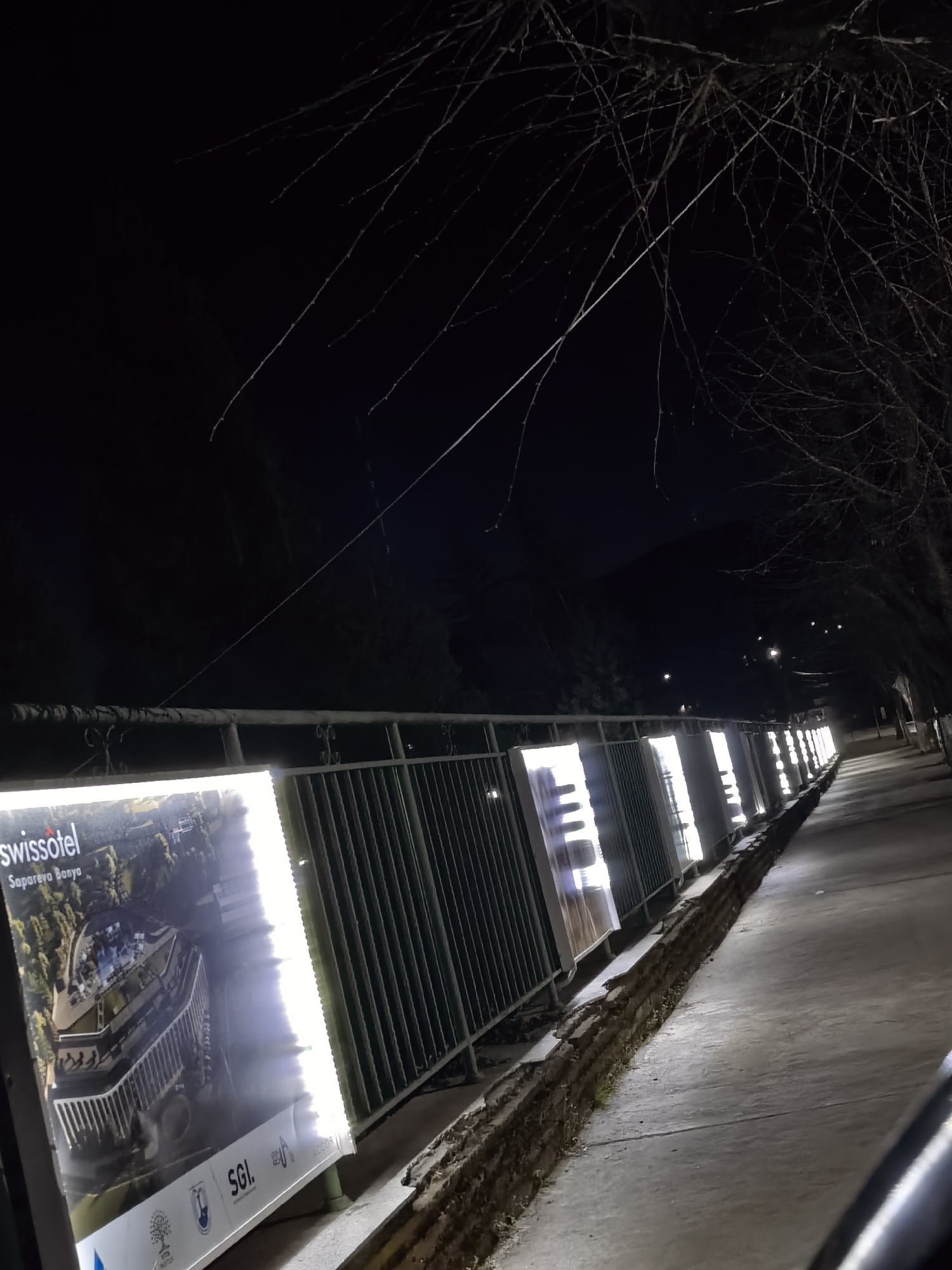 Captured at night, the scene features a line of illuminated signs. The signs are attached to a metal railing that runs parallel to a sidewalk. The signs display a promotional image for "swissotel Sapareva Banya", along with other logos, including "SGI". The railing and signs extend into the distance, illuminated by the signs' internal lighting. The sidewalk is grey and appears to be made of concrete. A low wall runs along the edge of the sidewalk, separating it from the railing. Behind the railing, there is a dark, wooded area. Overhead, tree branches are visible against the night sky. The location is Sapareva Banya in an unknown city.