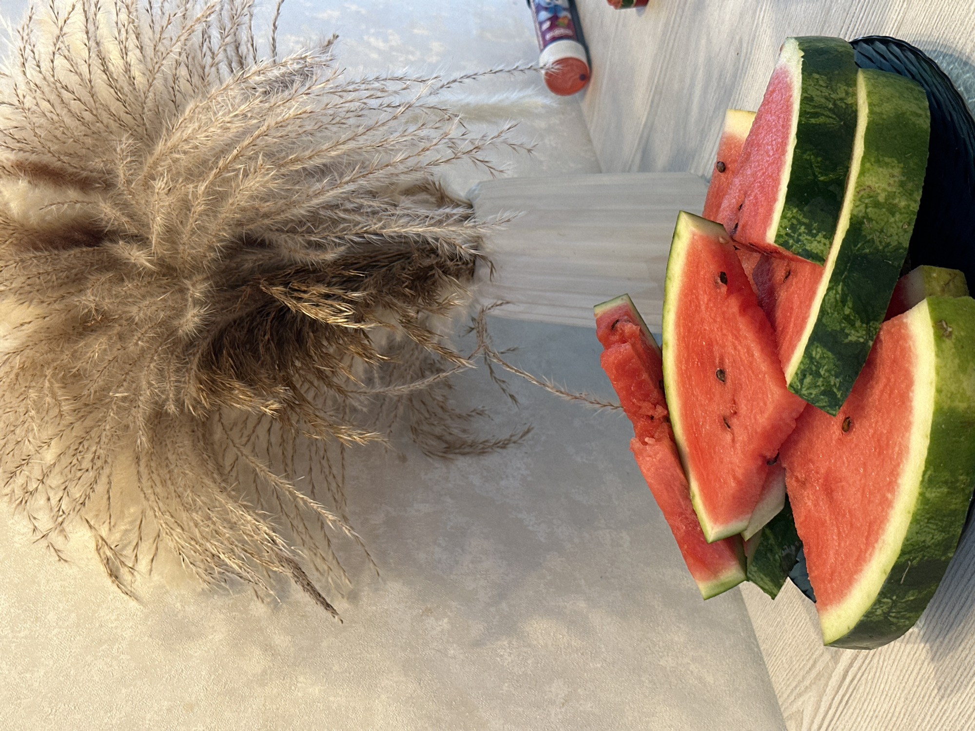 This media file shows slices of watermelon arranged in a dark bowl, next to a vase filled with dried pampas grass. The setting appears to be indoors, possibly on a table or countertop, with a textured, light-colored surface. The lighting suggests it could be daytime, with natural light casting shadows. The watermelon slices are triangular with visible seeds and the characteristic pink flesh and green rind. The dried grass is feathery and beige, creating a contrast in texture with the fruit. In the background, a white, stepped surface is visible, with a portion of what looks like a marker or pen. The overall impression is a still life arrangement, focusing on natural textures and colors. There is no visible text in the image.