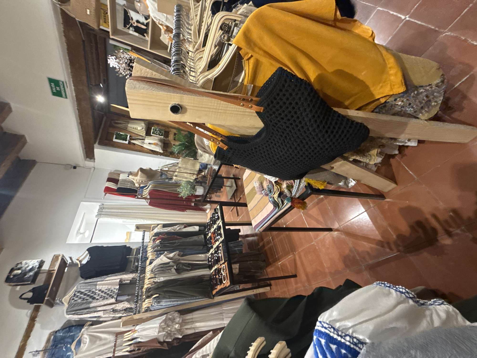 The image shows an indoor setting within a retail environment, likely a clothing or accessories store, in the city of San Cristóbal, Mexico. The floor is made of red-brown tiles. Clothing racks filled with various garments line the walls. A display table in the foreground presents sunglasses. A black crochet-style bag with brown leather straps is hanging on a wooden structure. A yellow garment is draped over a partially deconstructed chair or display. There are no people in the visible area.