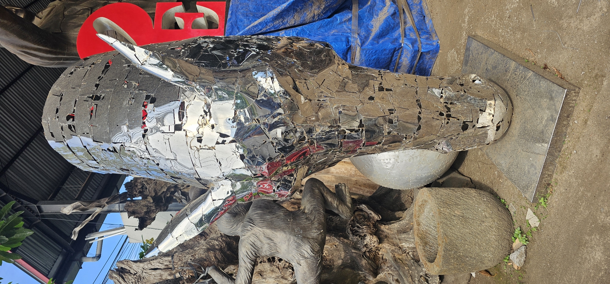 This image depicts a large metal sculpture of a horse's head in the Ubud District, Indonesia. The sculpture is constructed from small, irregularly shaped pieces of polished metal, creating a mosaic-like effect with numerous small openings throughout. The reflective surface of the metal pieces creates a fractured, distorted reflection of the surrounding environment. The horse head rests upon a metal square base. The sculpture is positioned on a concrete or dirt surface, surrounded by other objects. These include a large, carved wooden sculpture with the gray figure of an ape, a blue tarp, a half-sphere dome of silver metal with rivets, a large rock with a hole through it, and red letters, indicating that the sculpture is likely part of an artistic display or installation in an outdoor setting. The backdrop includes glimpses of a building with a corrugated metal roof and vegetation.