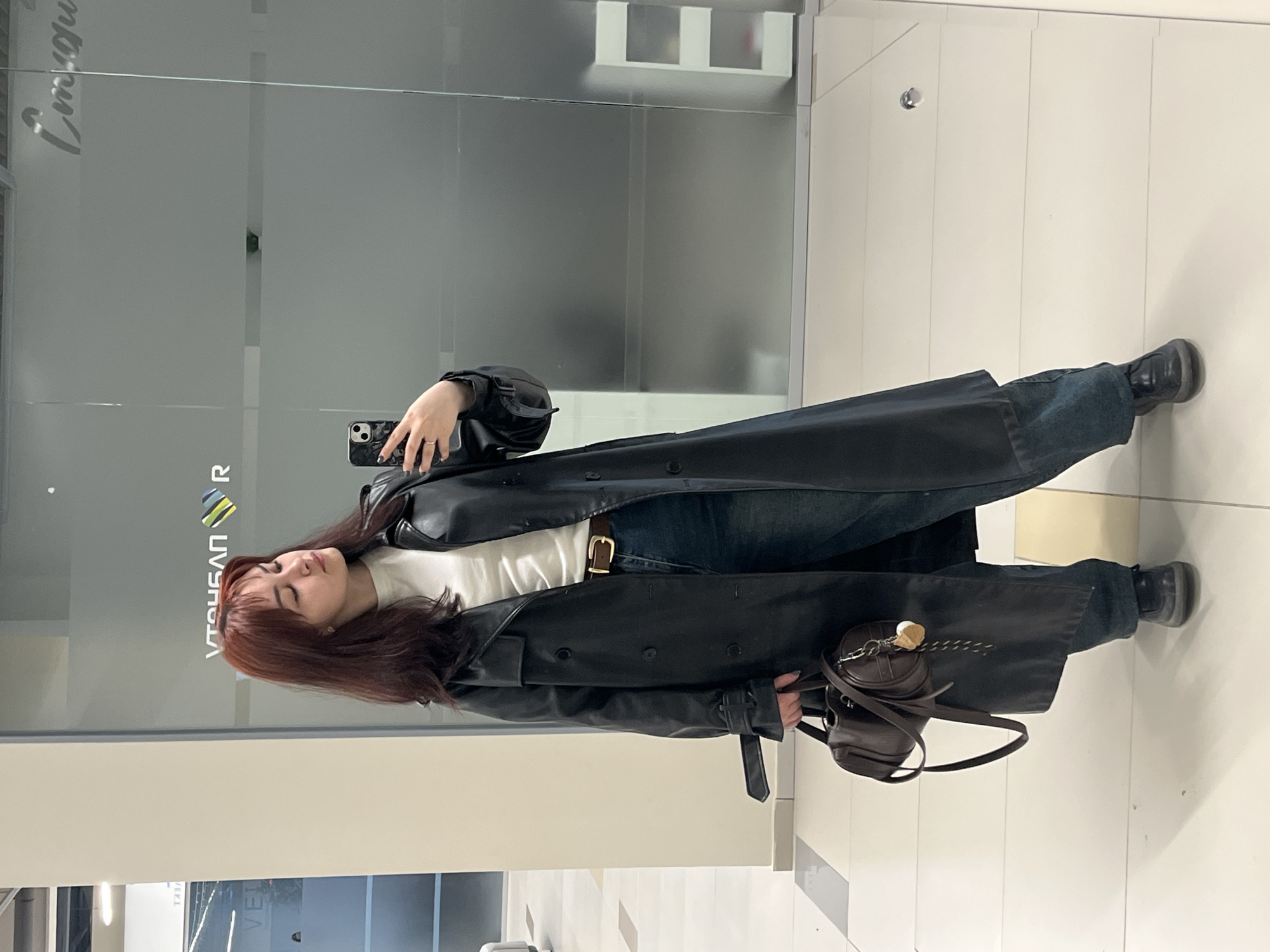 A woman is taking a selfie in a mirrored hallway. She is wearing a long black leather trench coat, a white shirt, and blue jeans. She is holding a cell phone in her right hand. A small brown purse hangs from her left shoulder. She is standing in front of a mirrored wall. The word "Neftekamsk" is visible on the glass. The floor is white tile. The location is assumed to be in the city of Neftekamsk, Russia.