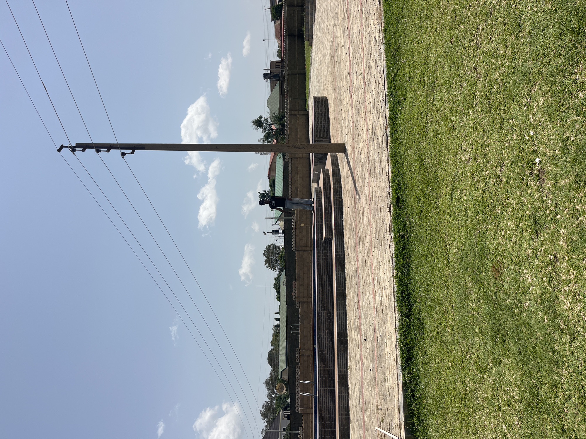 The image depicts an outdoor scene in Jos, Nigeria. The foreground features a paved walkway made of interlocking bricks and a strip of well-maintained green grass. A prominent utility pole with multiple wires stretches across the sky, indicating the presence of electrical infrastructure.
In the mid-ground, a person wearing a black t-shirt and light-colored pants is standing near the utility pole, facing away from the camera. They appear to be looking towards the buildings in the background. Behind the person, a low brick wall runs along the edge of the property, topped with a decorative element. Beyond the wall, several houses with distinct roofing styles and some trees are visible. The sky is bright blue with scattered white clouds, suggesting it is daytime, likely mid-morning or afternoon. The weather appears clear and sunny. The overall setting suggests a residential or possibly a communal area in Jos.