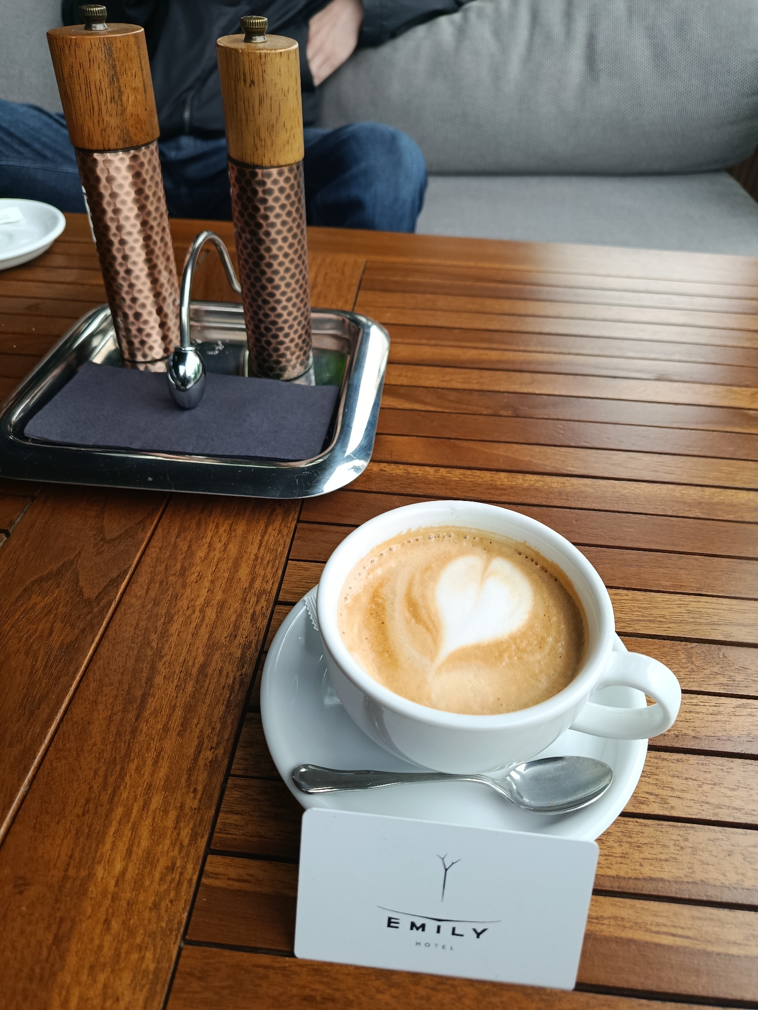 The image captures an indoor scene at a hotel or cafe in Vynnyky, Ukraine. The primary subject is a wooden slatted table, rendered in rich brown tones, extending from the foreground to the mid-ground.
On the right side of the table, a white ceramic cup holds a latte or cappuccino, adorned with a clear heart-shaped latte art in its foam. The cup rests on a matching white saucer, with a silver spoon positioned on the saucer. Directly in front of the cup and saucer, a white rectangular card is placed flat on the table, displaying the black text "EMILY HOTEL" beneath a stylized tree or branch logo.
On the left side of the table, a rectangular silver tray contains two cylindrical shakers. Each shaker features a light brown wooden top with a small brass finial, and a textured, hammered copper-colored body. Between the shakers, a small polished silver stand with a curved handle and a spherical base is visible. A dark gray or black rectangular napkin is folded beneath the shakers and stand on the tray.
In the soft-focus background, a person is seated on a light gray upholstered couch or bench. The individual is wearing blue denim jeans and a dark long-sleeved top or jacket, with one hand resting on their thigh. The background suggests a relaxed, well-lit interior environment.