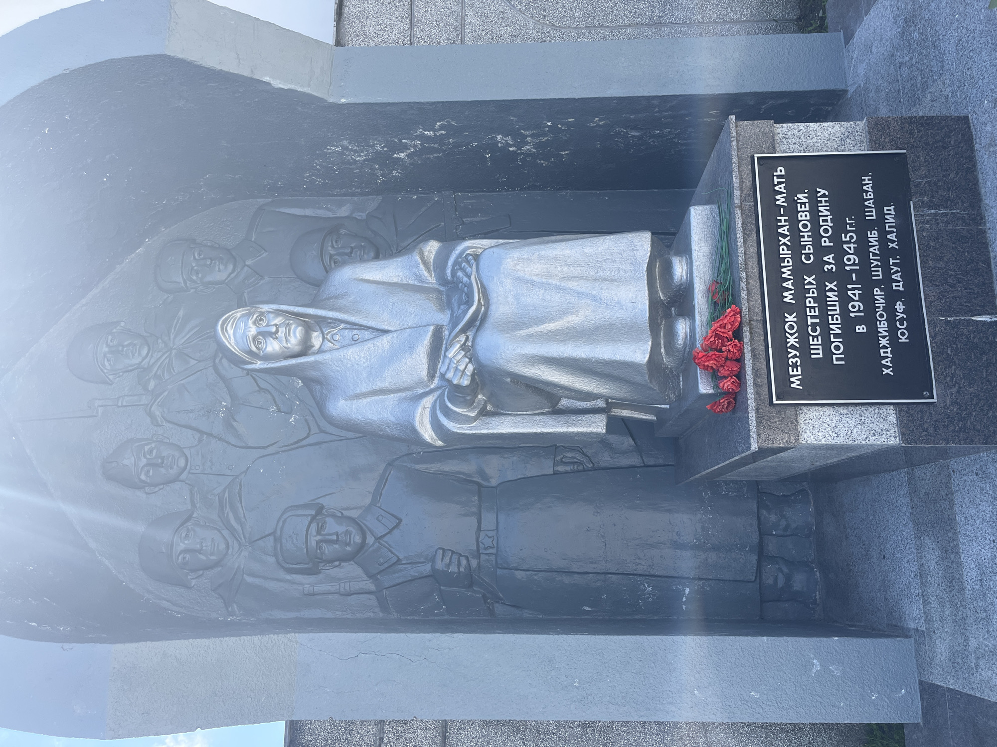 This media file captures a memorial monument dedicated to Mamyrkhan, a mother who lost six sons fighting for their homeland between 1941 and 1945. The monument features a central sculpted figure of a woman, depicted in relief, with a solemn and dignified expression. Surrounding her are the carved faces of men, likely her sons, also rendered in relief.
The monument is made of dark stone, with the sculpted figures appearing in a metallic silver finish. A black plaque with white Cyrillic text is affixed to the base of the monument, providing details about the sacrifice. The inscription reads: "METHYOK MAMYRKHAN-MATH / SIX SONS / WHO DIED FOR THE HOMELAND / IN 1941-1945 / KHAJIBOCHIR, SHUGAYB, SHABAN, / YUSUF, DAVUT, KHALID." A small bouquet of red flowers is placed at the base of the plaque, adding a touch of remembrance.
The monument is set against a backdrop of textured stone, possibly granite, with some areas appearing rough and others polished. The overall scene evokes a sense of solemnity, grief, and patriotic pride. The lighting suggests it was taken during the daytime, with some slight glare present. The location context indicates this memorial is in Takhtamukai, Russia, a place likely marked by historical events related to wartime sacrifices.