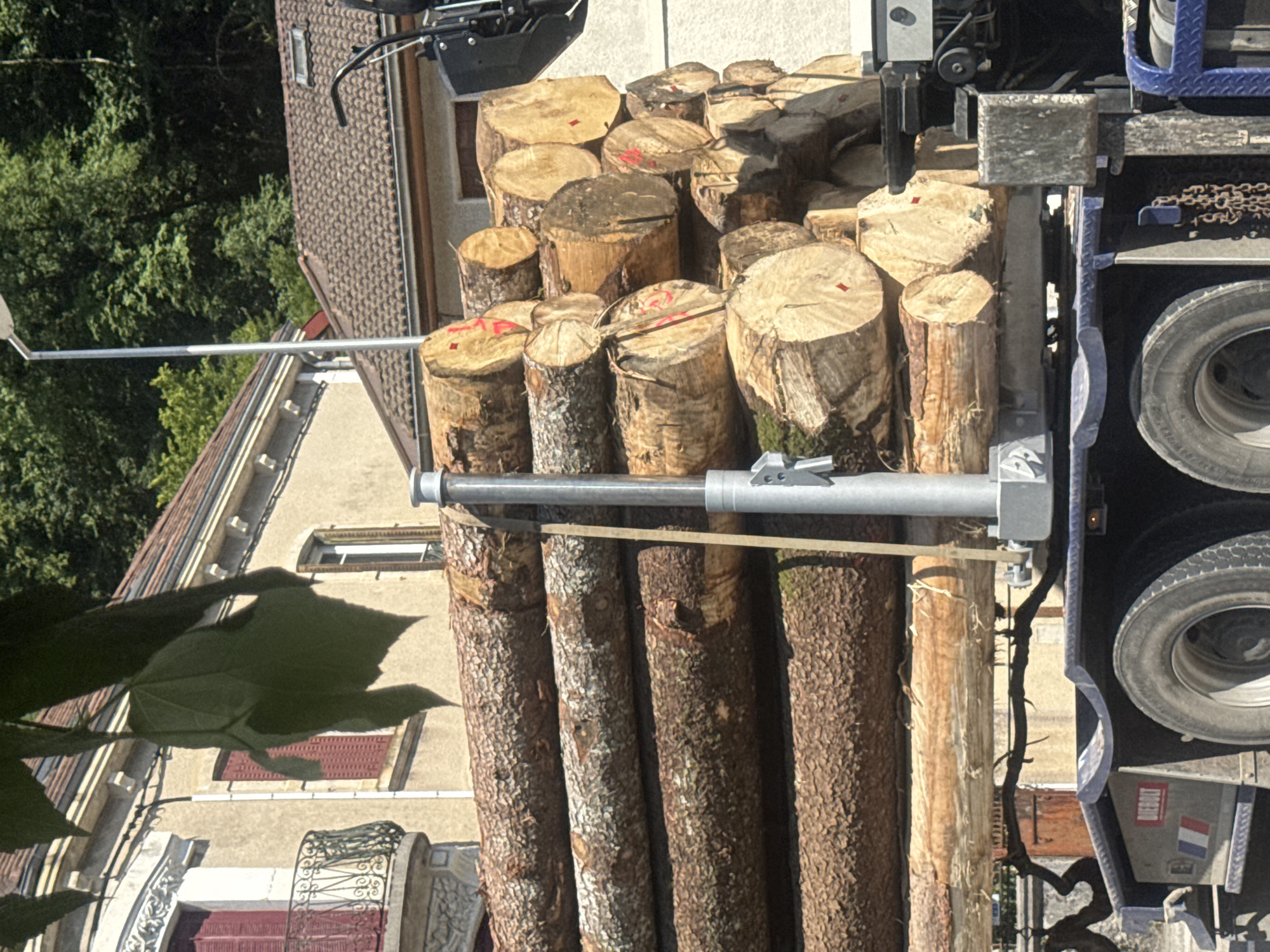 The image shows a close-up of a truck bed filled with logs. The logs are of various sizes and appear to be freshly cut, with some showing red markings on their ends. A metal arm or pole extends from the truck, crossing horizontally in front of the logs. The background includes parts of a building with a tiled roof and windows, as well as green trees. The lighting suggests it is daytime, likely sunny, given the strong shadows. The scene appears to be outdoors, possibly in a rural or semi-rural setting, consistent with a logging or construction site. There is some text visible on the truck, including what appears to be a flag sticker with blue, white, and red stripes, potentially indicating a French location. The overall impression is one of a work in progress, possibly related to construction or fuel preparation.