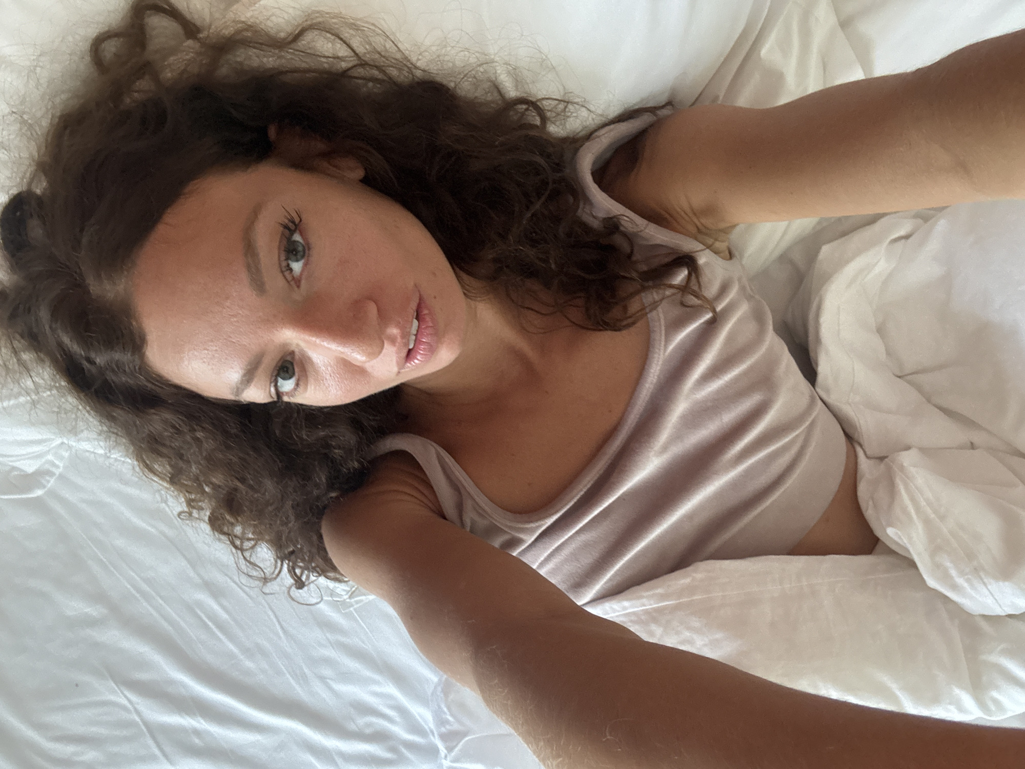 A woman with curly brown hair is lying in a white bed. She wears a light pink tank top. Her skin tone is fair, and she is looking at the camera. She is taking a selfie. The location in Đà Nẵng, Vietnam is not discernible from the image.