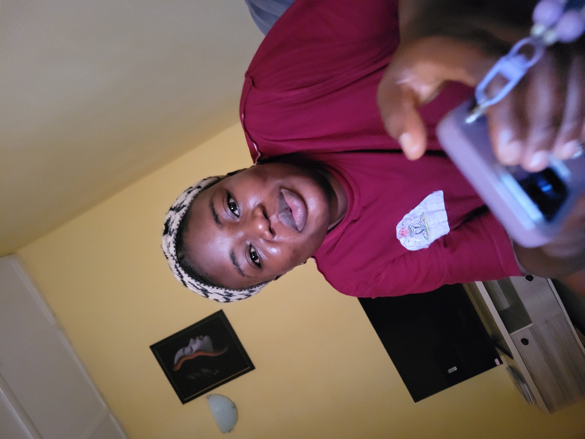 The image shows a medium shot of a young Black woman sitting indoors, likely in Suleja, Nigeria. She is wearing a maroon t-shirt and a patterned headscarf. The woman is looking directly at the camera, with her lips slightly parted. She is holding a light purple smartphone in her right hand, and a light purple lanyard is attached to the phone. A small patch with an emblem is visible on the left side of her t-shirt.
The background of the image is a light yellow wall. To the left of the woman, a framed picture of a woman's profile is hanging on the wall. Below the picture, a light fixture is mounted on the wall. In the lower left corner, a television set is visible on a wooden stand. The lighting in the room appears to be artificial, casting soft shadows. The overall impression is of a casual, indoor setting.