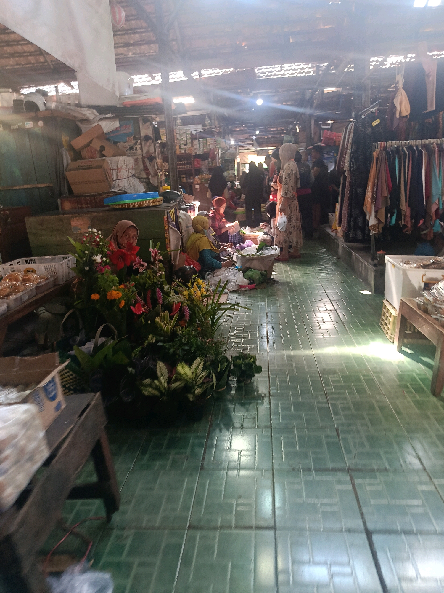 The image depicts a bustling indoor market scene, likely located in Yogyakarta, Indonesia, given the visual cues such as the presence of Islamic clothing and the overall market atmosphere common in the region. The foreground features stalls displaying a variety of goods. On the left, a stall presents fresh flowers, with a woman wearing a hijab tending to them. Next to the flowers, there are baskets filled with what appear to be baked goods or pastries. Further back, a clothing stall is visible with racks of colorful garments.
The market floor is covered in green tiles, with sunlight streaming in from above, creating bright patches of light and shadows. Several people are scattered throughout the market, some appearing to be vendors and others customers. Their attire suggests a conservative cultural context, with many women wearing hijabs and modest clothing. The background shows the market's structure with a wooden roof and various items stacked on shelves or in boxes, indicating a typical marketplace setting. The overall impression is one of lively commerce and daily life unfolding within the covered market space.