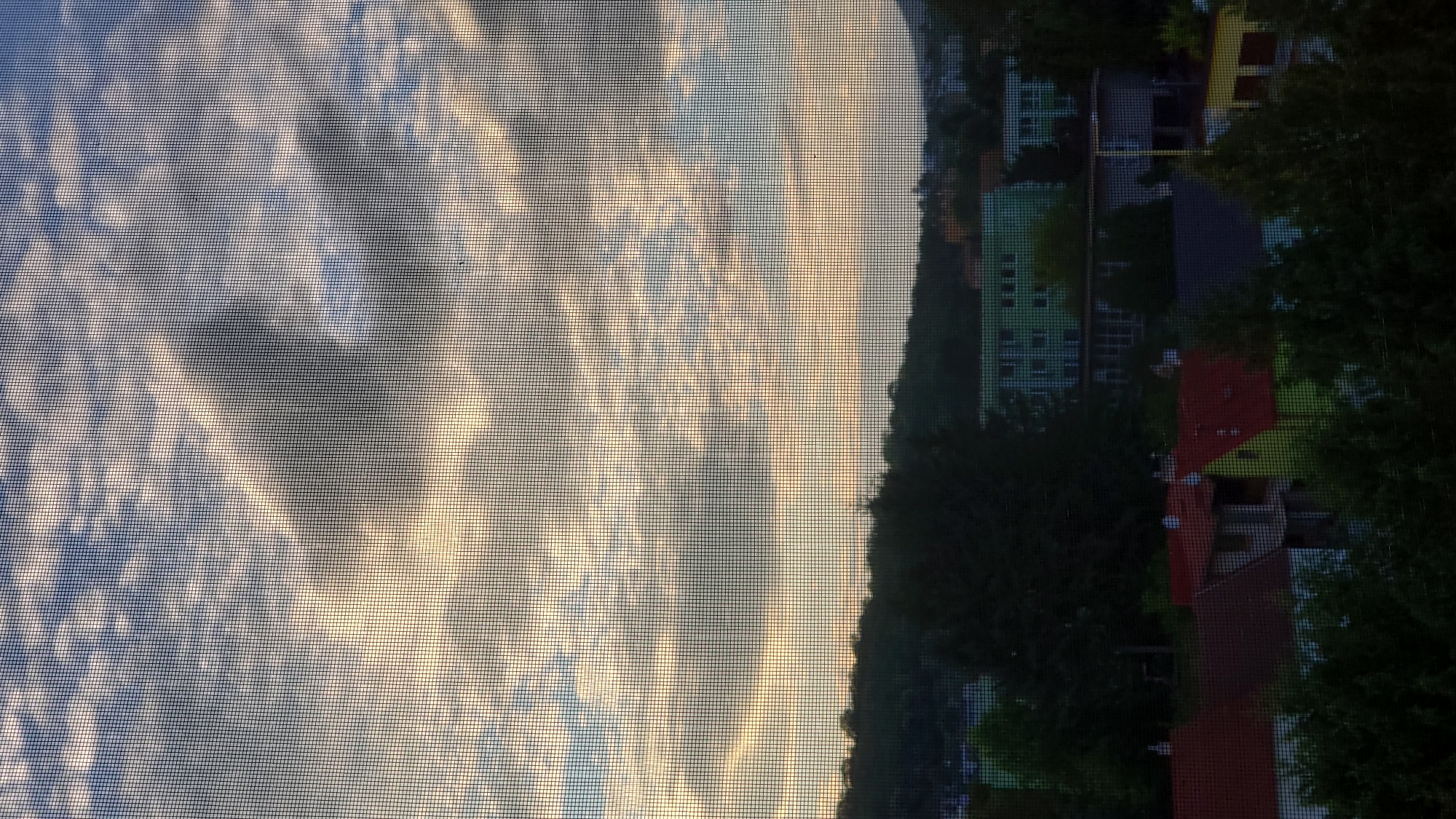 The image shows a view of a residential area in Prešov, Slovakia, seen through a window screen. The sky is partly cloudy with sunlight breaking through, casting a warm glow on the scene. Below the sky, a tree-covered hillside forms a dark silhouette against the lighter sky. In the middle ground, several buildings are visible, including a multi-story green building and a longer, lower building with a dark facade. In the foreground, various houses with red and yellow roofs are partially obscured by trees. The overall impression is of a suburban or semi-urban landscape during daylight hours, likely late afternoon or early morning given the quality of light.