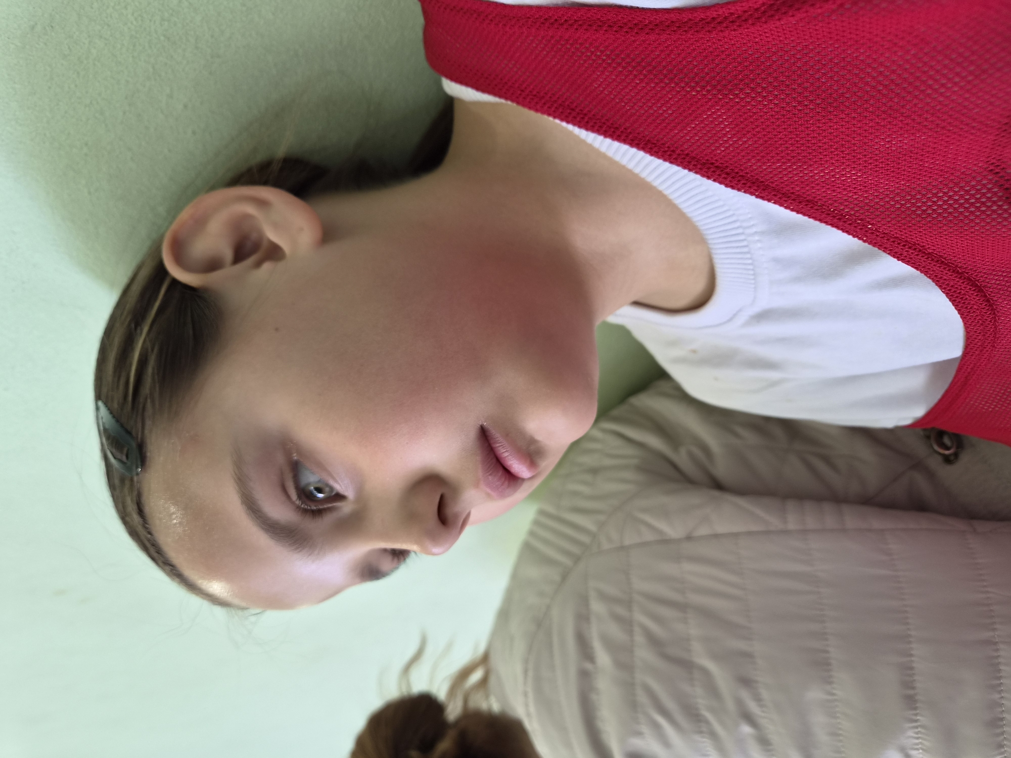 A young person is shown in a close-up, angled shot. They have fair skin, light brown hair with a dark hair clip, and are wearing a white shirt with a red mesh vest on top of what appears to be a tan-colored quilted jacket. The background is a light green color. This photo was taken in the city of Chaikovsky, Russia.
