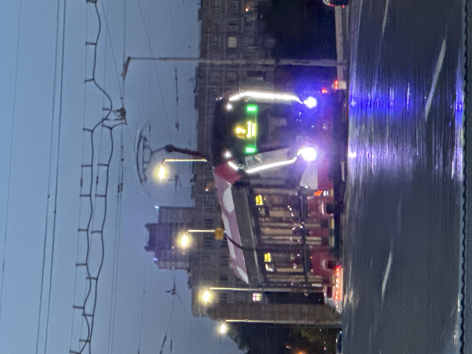 A street-level, eye-level, full shot of a red and white tram moving towards the viewer at dusk in Kudrovo, Russia. The tram has bright white and green lights on its front, with a yellow number "7" displayed in the center. The tram is reflected in the wet asphalt road, which is illuminated by the tram's headlights and other streetlights. In the background, there are several multi-story buildings and overhead power lines. The sky is overcast and a dim blue.
