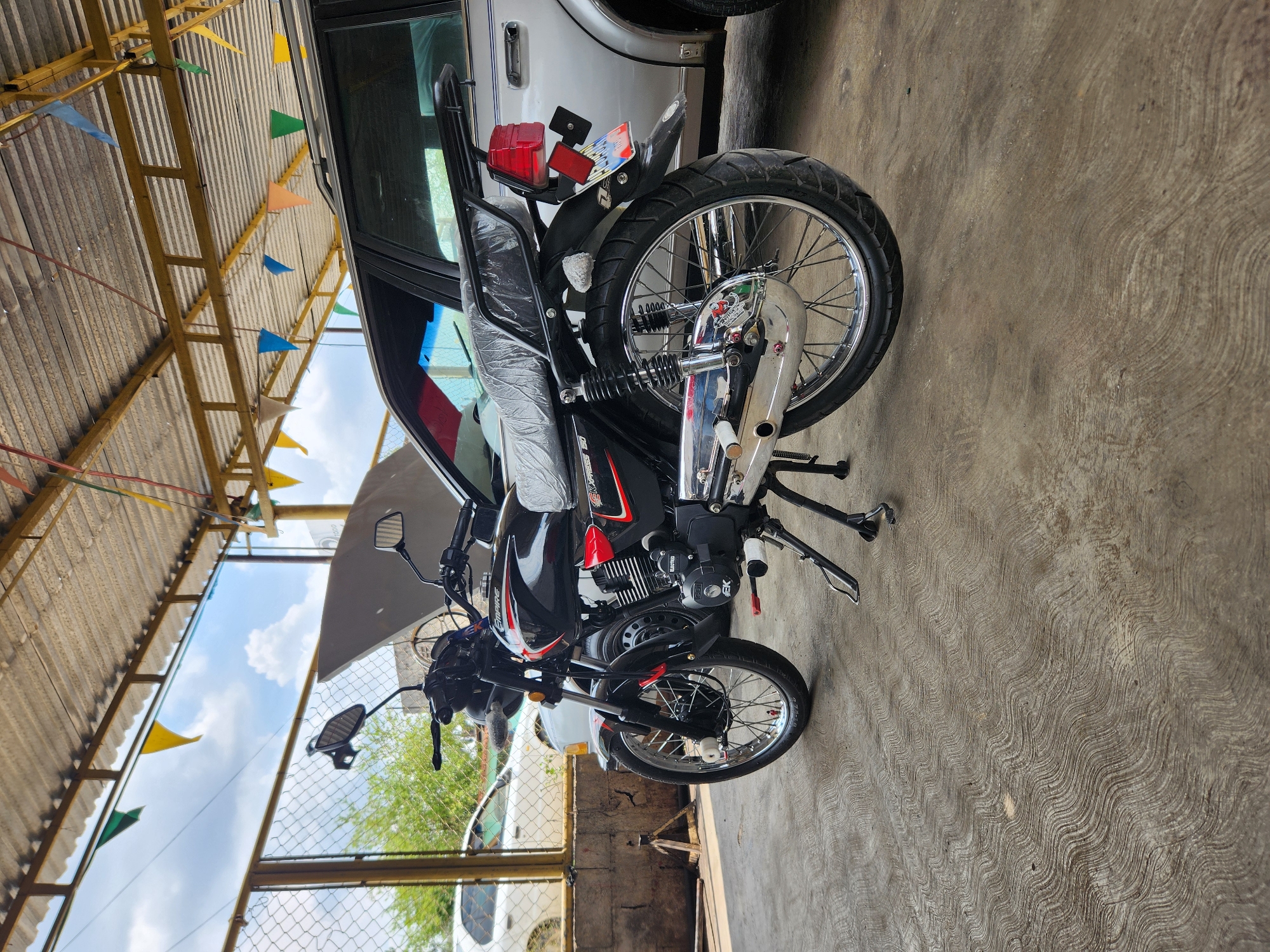 This image captures a black motorcycle parked indoors, likely in a garage, carport, or open-air service area, next to a white car. The photo is taken with a vertical orientation, making the ground appear on the right side and the roof structure on the left.
The central subject is a black motorcycle with red and white accents and decals, including "EMPIRE" visible on the fuel tank and "EXPRESS 150" (or similar) along with "GK" visible on the engine or side cover. The motorcycle's seat is covered in clear plastic wrapping, suggesting it is new, recently purchased, or has just undergone servicing. It features chrome spoke wheels, a disc brake on the rear wheel, and a black metal backrest or luggage rack behind the seat. The rear fender holds a red taillight assembly with what appears to be a temporary license plate or dealer tag, flanked by two red rectangular reflectors. The motorcycle is standing upright on its kickstand.
To the immediate left of the motorcycle, a portion of a white car is visible, specifically its side and rear. The car appears to be an older model sedan or SUV.
Above the motorcycle and car, a corrugated metal roof structure is visible, supported by yellow-painted metal beams and trusses. Strung across these beams are numerous small, triangular flags in various colors including blue, yellow, green, orange, and red, adding a festive touch to the setting.
The ground beneath the vehicles is a concrete slab with distinctive, wavy, textured patterns. On the far left of the image, a chain-link fence is visible, and beyond it, some green foliage (likely trees or bushes) can be seen. Through gaps in the roof structure and above the fence, a bright blue sky with white clouds is visible, indicating it is daytime. The scene is well-lit, suggesting either morning or afternoon lighting.