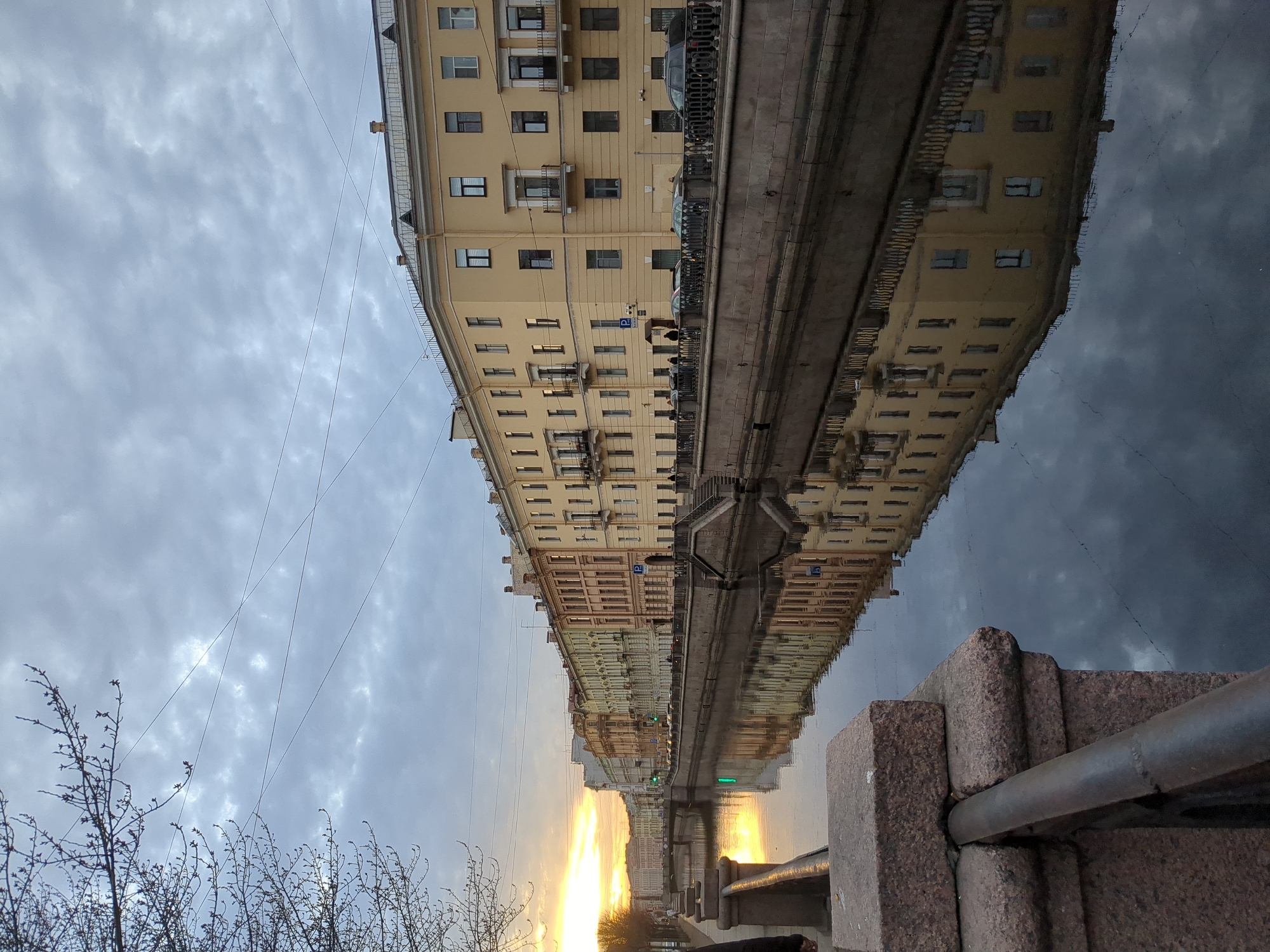The image captures a canal scene in Saint Petersburg, Russia, likely taken near the city center due to the architecture and canal presence. The shot is composed with the canal and its reflection dominating the frame. Buildings flank the canal, their facades reflecting in the water. The buildings are of varying heights and colors, with a mix of pastel hues. A series of parked cars can be seen along the canal's edge on the side adjacent to the buildings. The sky is overcast with scattered clouds, and a warm, golden light suggests either sunrise or sunset. A granite embankment with a metal railing is visible in the foreground, indicating the position of the photographer. There are some bare branches of a tree visible on the left side of the frame. There are no people visible in the image.
