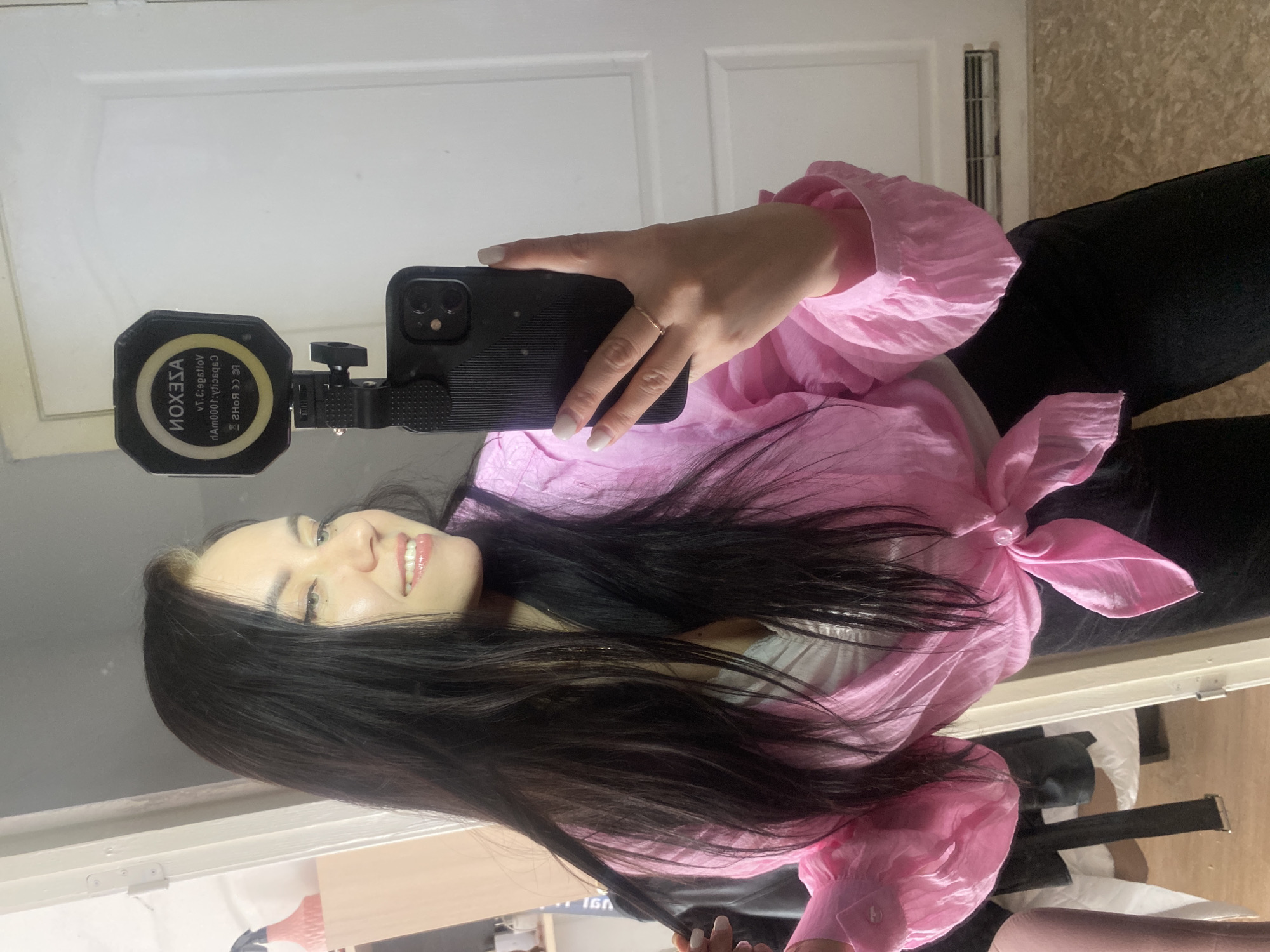 An image shows a young woman taking a mirror selfie indoors, with the photo rotated 90 degrees counter-clockwise. She is smiling broadly, revealing her teeth, and has long, dark hair with some lighter streaks near her face. She wears a light pink, flowy long-sleeved blouse, tied at the front, and dark pants. Her nails are painted a light color, and she wears a thin ring on her right ring finger.
In her right hand, she holds a black smartphone, featuring a dual-camera setup, with a small, octagonal black LED light attached, which has a distinct yellow ring. The LED light displays mirrored text: "AXESON" at the top. Below this, also mirrored, are technical specifications: "VOLTAGE: S.T.V", "OUTPUT: 1000MA", and certifications such as "CE ROHS2 CE FC".
The background, visible in the mirror's reflection, includes a white door or wall, a vertical vent, and a section of what appears to be textured wallpaper or another wall covering. The scene depicts a casual indoor setting. While the location context is Chaikovsky, Russia, no specific visual cues within the image confirm this.