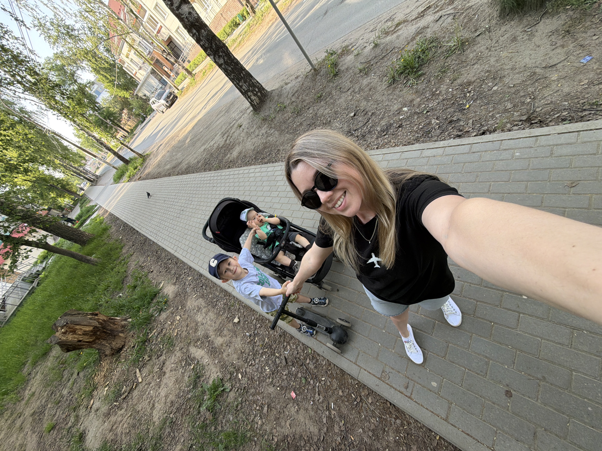 A woman is taking a selfie on a brick paved sidewalk in Chaikovsky, Russia. She is wearing sunglasses, a black shirt with a white airplane on it, and white sneakers. Next to her is a black stroller with a baby inside. In front of the stroller, a young boy stands on a black scooter and holds the woman's hand. To the left of the sidewalk is a dirt area with patches of grass and a tree stump. Behind the sidewalk is a road, trees, and buildings. A small bird sits on the sidewalk ahead of them.