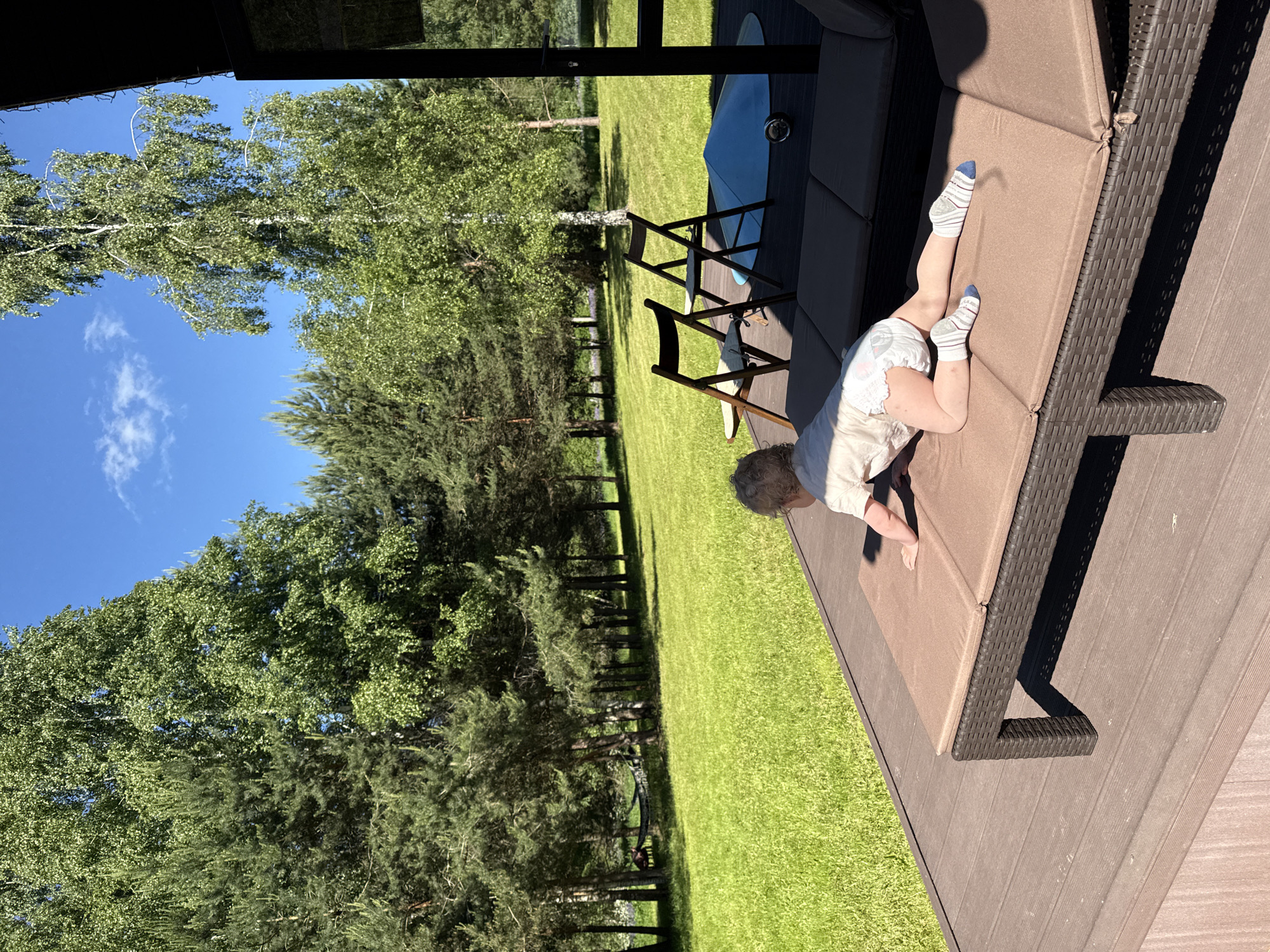 The image depicts an outdoor scene in Копнино, Russia, under bright daylight with a clear blue sky. In the foreground, a young child, appearing to be a toddler, is positioned on their stomach on a dark brown wicker-style sun lounger with light brown cushions. The child has light-colored curly hair and is wearing a light-colored short-sleeved shirt, a diaper, and white socks with blue and gray stripes. Marks are visible on the child's legs. The child's arms are extended forward, supporting the upper body, while the knees are bent, suggesting a crawling or climbing motion. The lounger is situated on a brown slatted deck or terrace, which occupies the lower portion of the frame. To the right of the lounger, two dark wood folding chairs with light cushions are visible, facing outward towards the lawn. Further to the right on the deck, a light blue conical object, possibly a recreational item, and a small metallic sphere are present. A dark structure, likely part of a building, is partially visible on the far right edge of the frame.
Beyond the deck, an expansive, well-maintained green lawn fills the midground. In the background, a dense line of various trees borders the lawn. These include numerous pine trees (conifers) on the left side and several birch trees (deciduous with light bark) scattered throughout, particularly prominent in the center-right, all with full green foliage. A dark-colored hammock is visible strung between two trees in the distant left background. The sky above is a clear blue with a few small white clouds. Strong shadows indicate a high sun position.