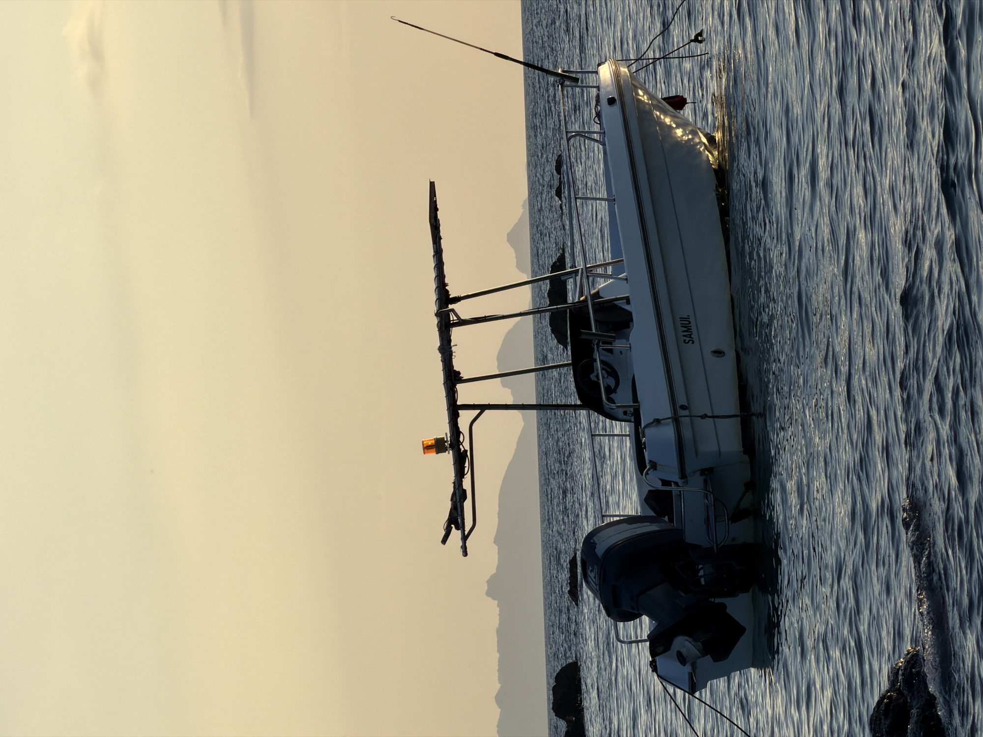 A white motorboat with an outboard engine is anchored in calm, dark blue water near the shore. The boat has a canopy supported by metal poles and an orange flashing light on top. A fishing rod is mounted at the stern. The words "SAMUI" are visible on the side of the hull. In the background, silhouetted mountains rise from the sea under a pale yellow sky, suggesting a sunset or sunrise. Rocks are visible in the water near the boat and in the foreground. The scene is located in the sea off the coast of Ko Samui District, Thailand.