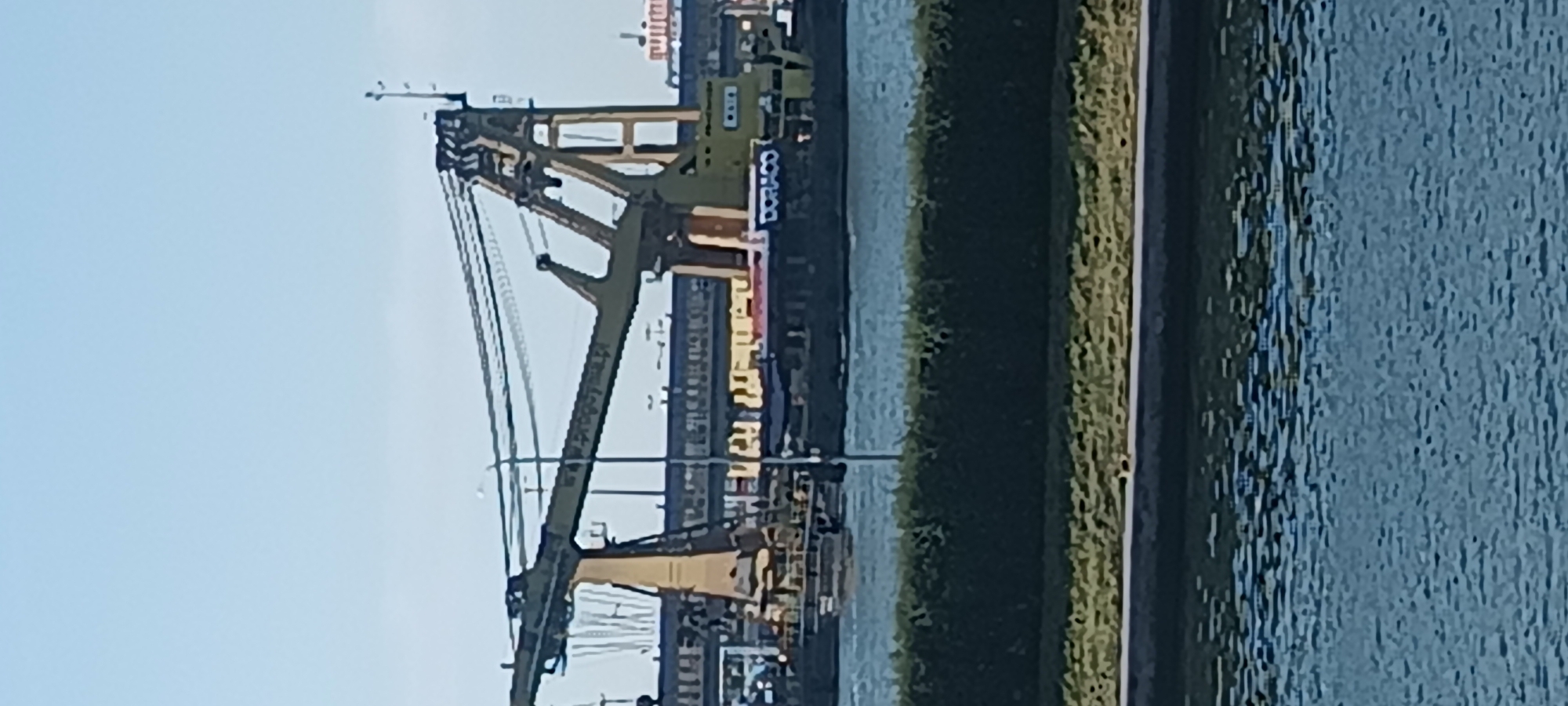 An outdoor, daytime, medium-wide shot captures a large yellow crane on a barge in the foreground. The crane's boom extends across the frame, supported by a tall A-frame structure. In the background, a long building with a yellow roof runs horizontally. The building appears to be part of an industrial complex, possibly a shipyard or port facility, located in the city of Świnoujście, Poland. A blue body of water is visible in the lower portion of the image, reflecting the sky and surrounding structures. The scene is dominated by the industrial infrastructure and the calm water, suggesting a working harbor environment.