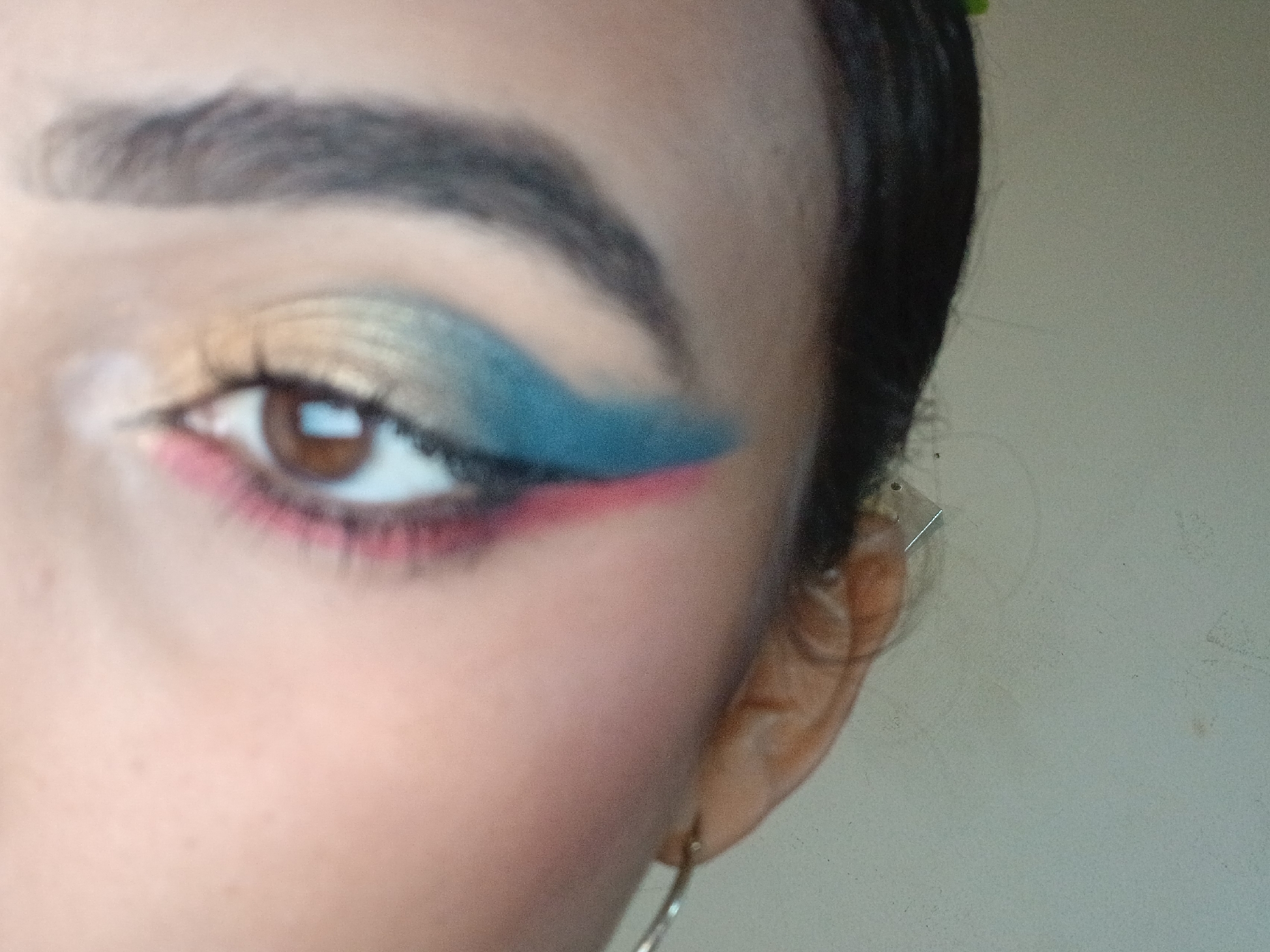 The image depicts a close-up of a person's eye and a portion of their face. The person has an eyebrow shaped with makeup, and their eye is adorned with colorful eyeshadow in shades of yellow, green, teal, and red. A hoop earring is visible on their ear. The background is blurry, with a hint of a light-colored wall. There is nothing in the picture to suggest the location of Playa El Agua, Venezuela.