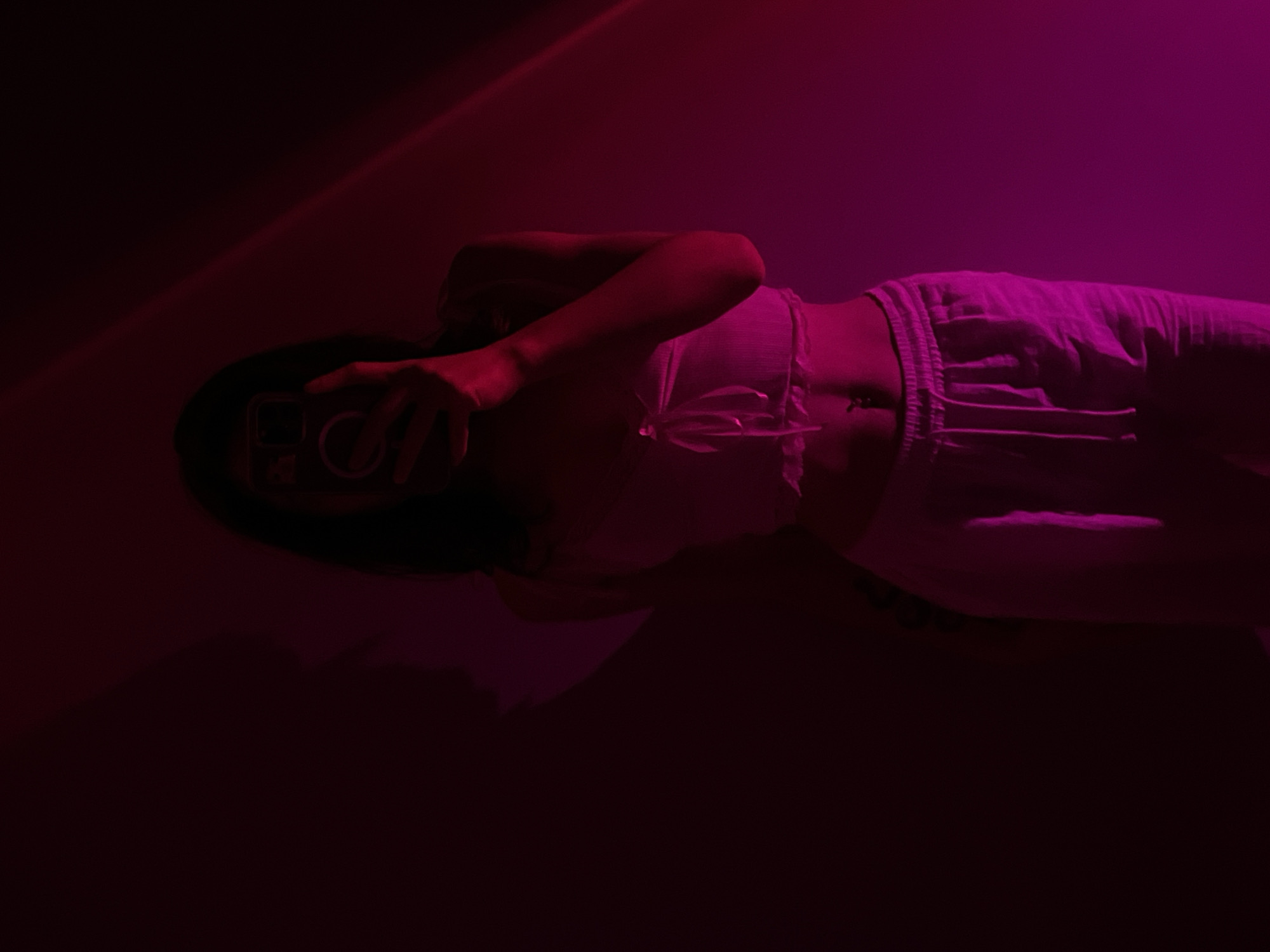 A full-body mirror selfie of a person is taken in a dimly lit room with pink and red lighting. The person is wearing a light-colored crop top and light-colored sweatpants. They are holding a smartphone up to take the picture. The background is dark, with a shadow cast on the left side of the image. There is a light beam across the top right of the image. There is no information to suggest the location is Ternopil, Ukraine.