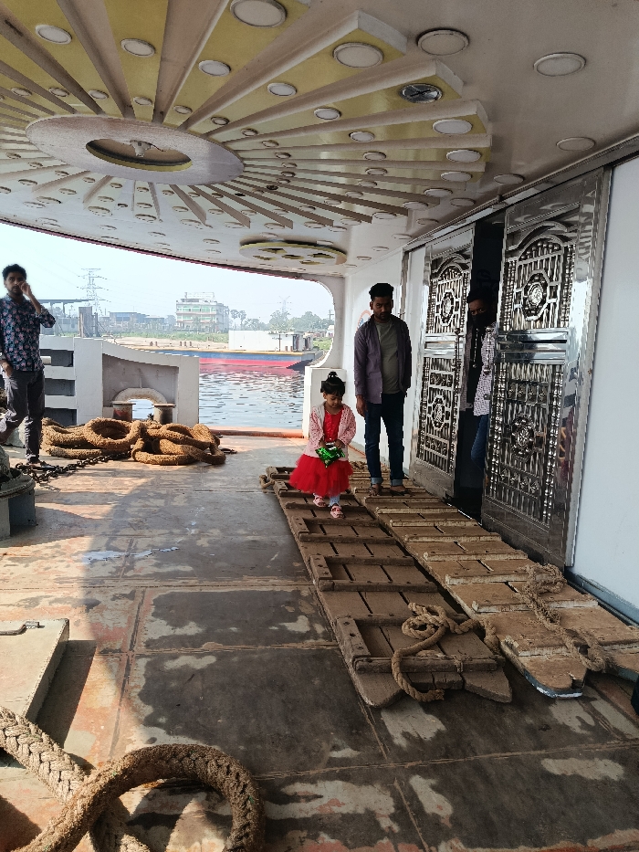 The image depicts a bright, sunny scene on the deck of a boat or ferry, likely in Zazira, Bangladesh. The time of day appears to be midday or early afternoon, indicated by strong shadows and clear skies.
In the foreground, a small child, dressed in a vibrant red frock with a white underskirt, stands on two rustic wooden ramps placed on the deck. She holds a green, possibly wrapped, item in her hands and looks downwards. To her immediate right, an adult male wearing a purple-grey long-sleeved shirt and dark jeans stands, looking towards the right of the frame. On the far left, another adult male in a patterned dark shirt stands near large coiled ropes, with his hand near his face. A third adult male is partially visible on the far right, standing near a set of ornate, polished silver metal doors that are slightly open.
The boat's deck is expansive and appears weathered, with visible metal plates and dark stains. Several thick, brown ropes are coiled on the left side of the deck, suggesting mooring or operational equipment. The ceiling above is intricately designed with a central circular light and radiating panels embedded with numerous smaller circular lights, forming a decorative sunburst pattern.
Beyond the boat's edge, a wide river stretches into the background. A large red and blue barge or boat is visible further along the river. The distant riverbank features various buildings and a tall electricity pylon, indicating a populated, possibly industrial or semi-urban, area. The scene captures a casual moment of individuals on a vessel in a riverine environment. No visible text is present.