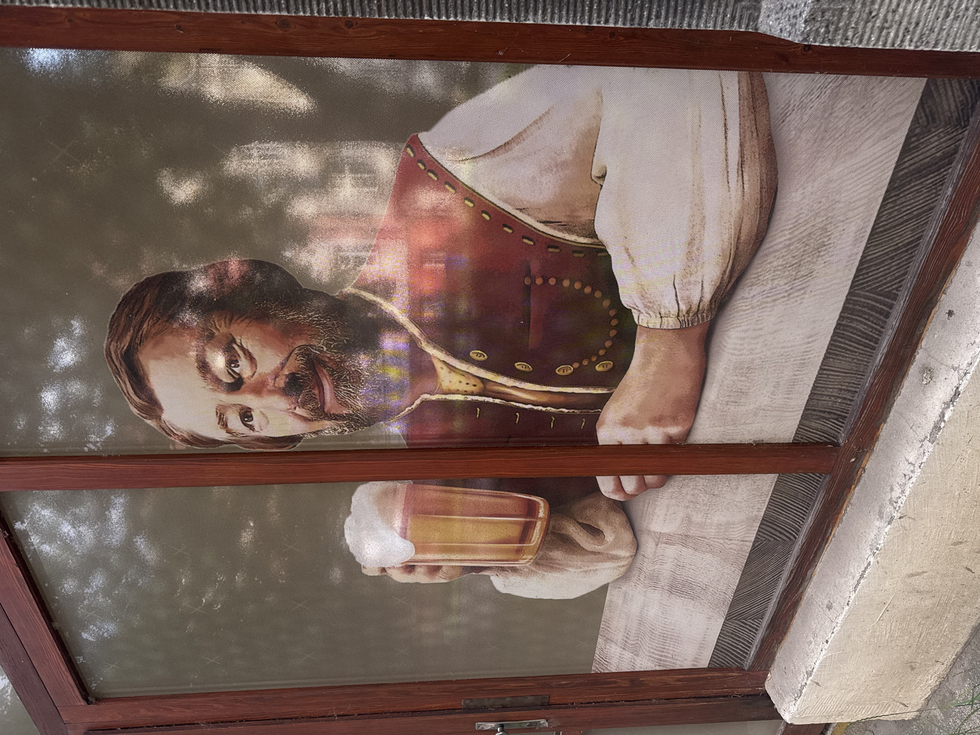 This is a medium shot photograph of a poster on a window, likely at an establishment in Prostějov, Czechia. The poster depicts a man with a beard, wearing a traditional vest over a white shirt, holding a large glass of beer. He is leaning on a wooden surface. The background of the poster shows a blurry outdoor scene with trees. The window has a wooden frame and is set into a concrete wall. A metal handle is visible on the left side of the frame.