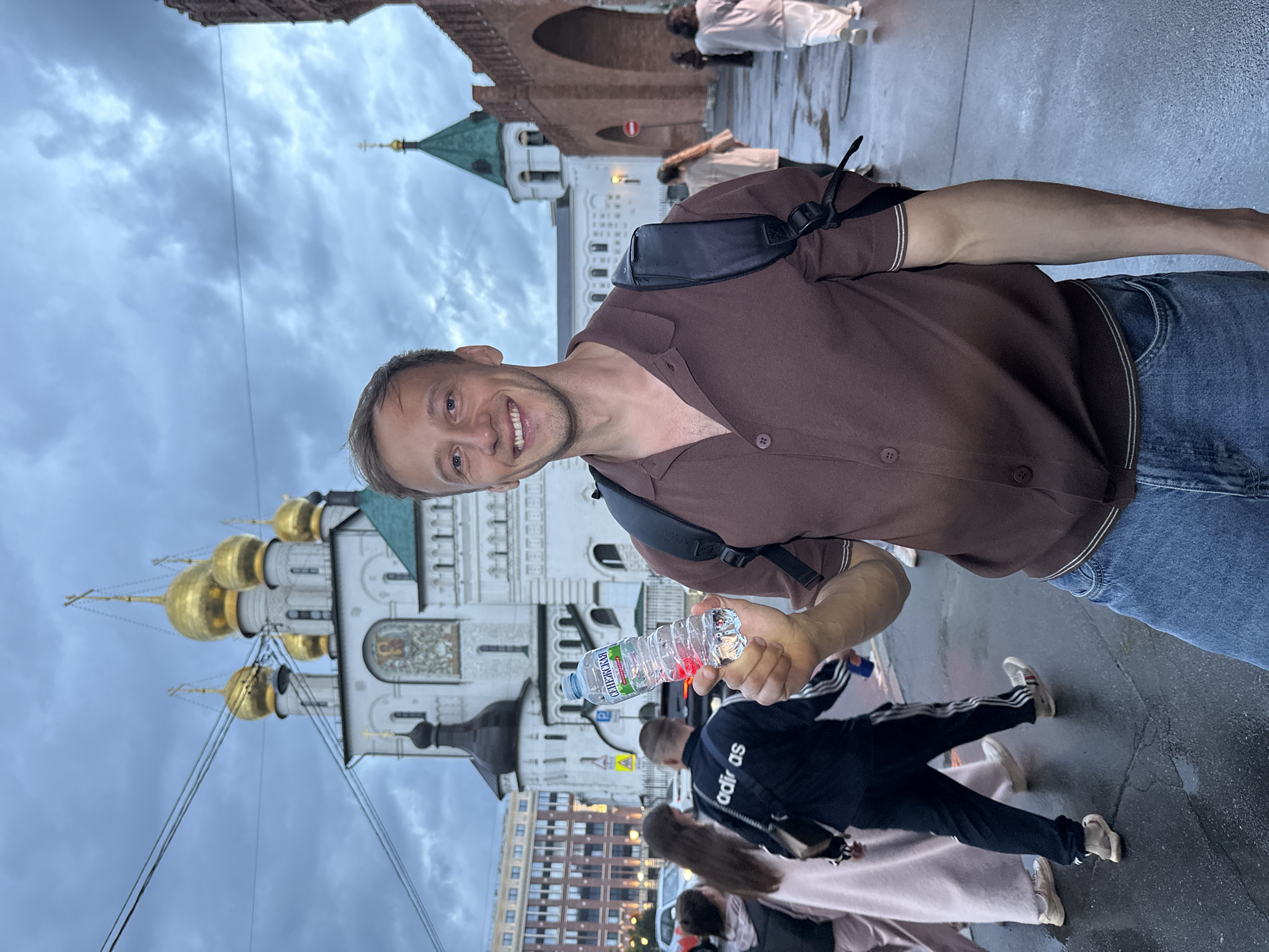 A smiling man with short brown hair and blue eyes is holding a plastic bottle of water in his right hand. He is wearing a brown collared shirt with dark trim and blue jeans. A black backpack is slung over his shoulders. In the background, a white church with golden domes and a green-roofed tower is visible. A section of a red brick wall with an arched gateway is to the right. Several people are walking in the wet, paved area in front of the buildings. The sky is overcast and grey. The man is in the foreground, positioned slightly off-center to the right.