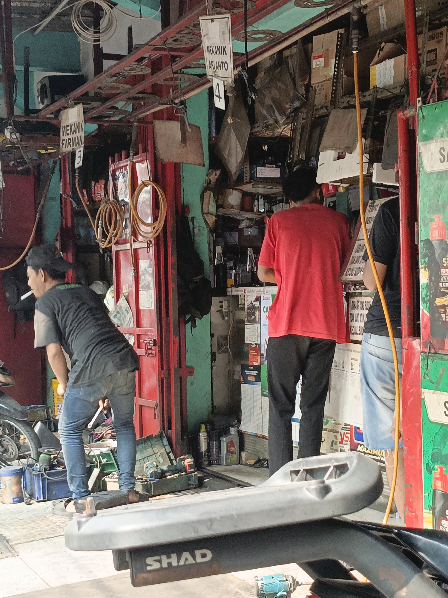 The image depicts a street-side motorcycle repair shop in East Jakarta, Indonesia. The scene is cluttered with tools, spare parts, and various workshop items. In the foreground, a partially visible gray motorcycle part with "SHAD" branding is prominent.
Three individuals are present. The person on the left, wearing a black t-shirt, dark pants, and a black cap, is smoking a cigarette and appears to be leaning over, possibly working on a motorcycle. In the center, a person in a red t-shirt and black pants stands facing away from the camera, seemingly engaged in activity within the shop. To the right, a person wearing a dark t-shirt and blue denim shorts is visible from the waist down, also facing away.
The shop's interior is characterized by metal shelving laden with boxes and items. Red and teal painted walls form the backdrop. Hanging from the ceiling are coiled hoses and electrical wires. Signs in Indonesian, such as "MEKANIK FIRMAN 3" and "MEKANIK ARI ANTO 4," indicate the presence of mechanics and their assigned numbers. Various tools, including a blue impact wrench, are scattered on the ground. Oil cans and other workshop supplies are visible on shelves and the floor. The overall impression is that of a functional, albeit busy and somewhat disorganized, automotive repair establishment in a bustling urban environment.