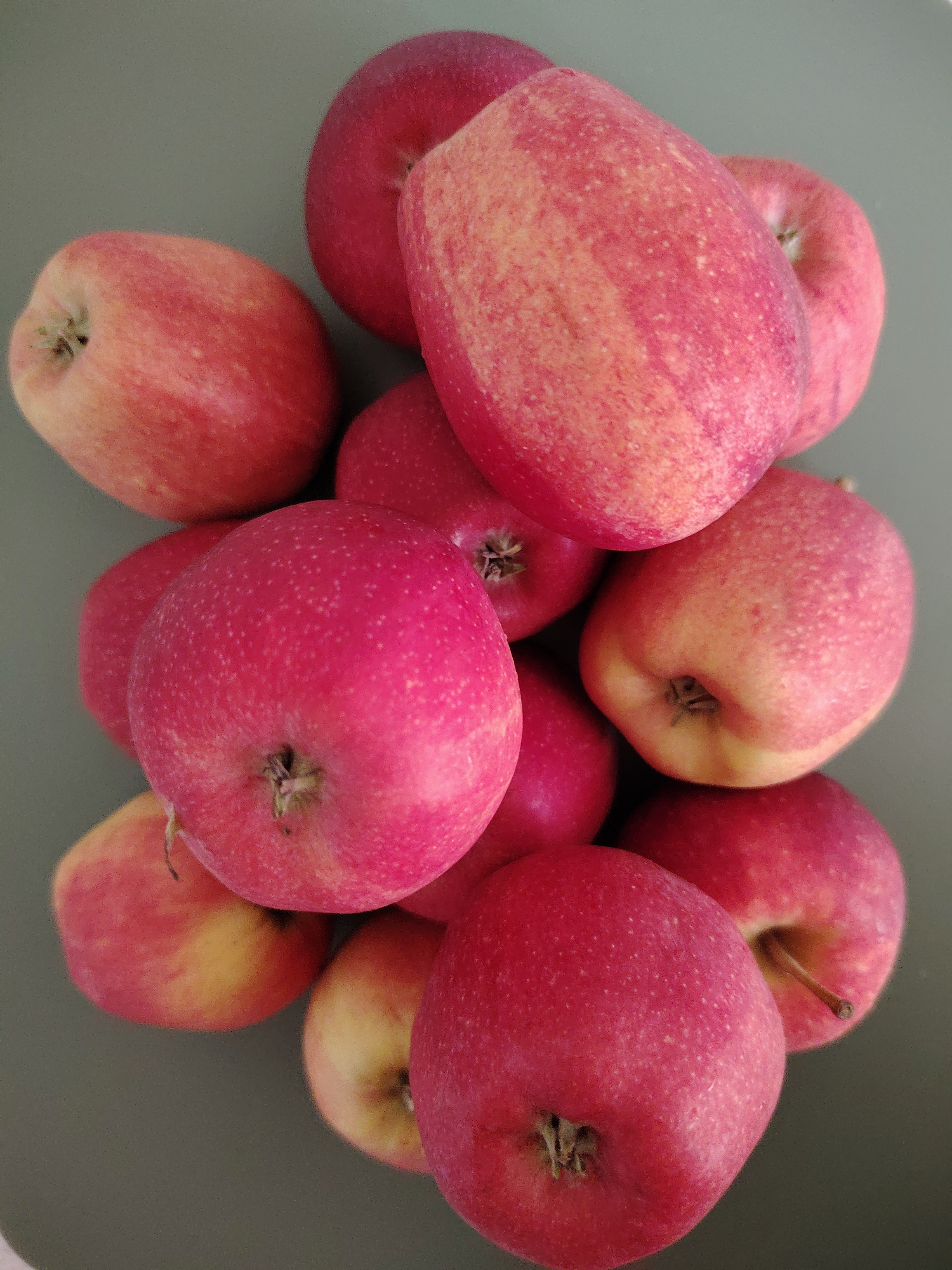 A close-up, top-down shot shows a pile of approximately ten bright red apples with some yellow and green undertones, clustered on a plain, muted green surface. The apples vary in size and are arranged in a seemingly haphazard pile, with some partially obscuring others. Their stems are visible on a few of the apples. The lighting casts subtle shadows, highlighting the roundness of the fruit. The image is sharp and focuses on the texture and color of the apples. There are no people or other objects visible in the frame. The location in Кривец, Russia is not discernible from the visual information provided in the image.